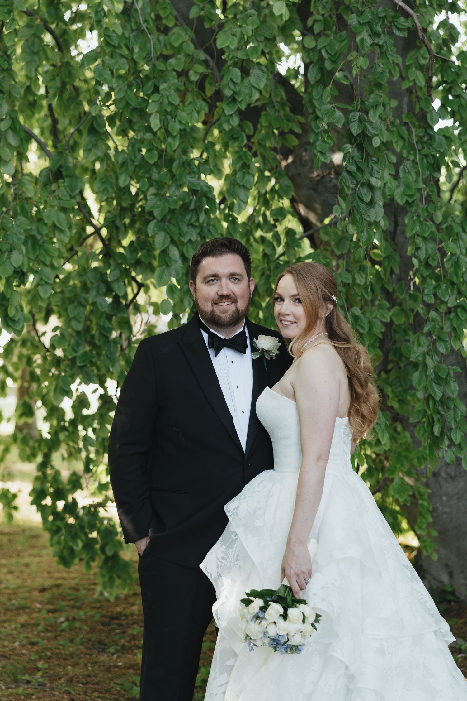 A bride and groom pose together under a lush green tree, she holds a white bouquet in her layered wedding gown.