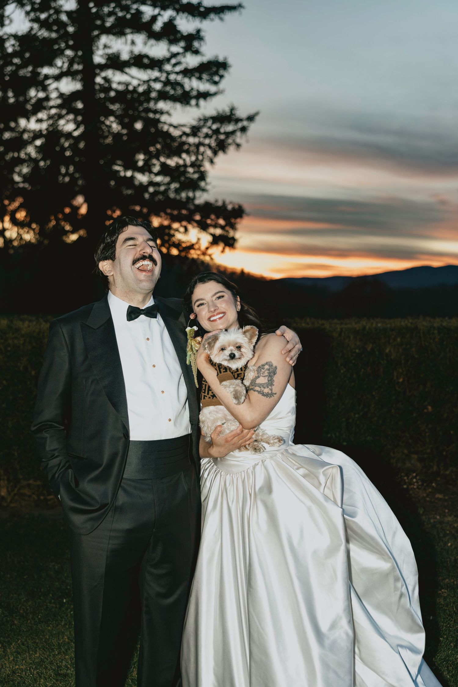 Couple in formal wedding attire laughs together while holding their small dog at sunset with mountains in background.
