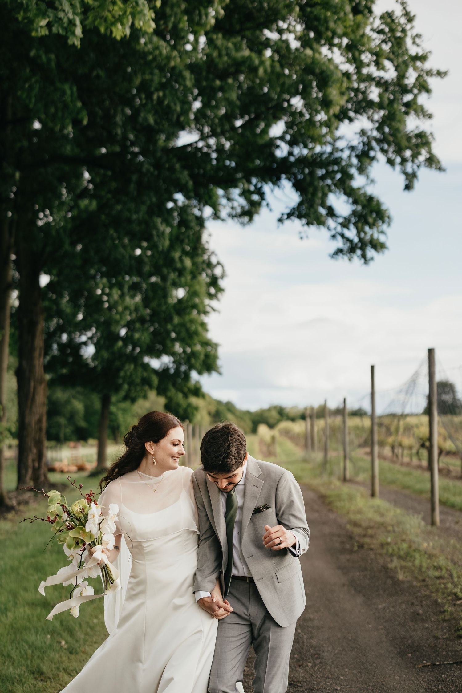 A couple shares a joyful moment walking along a tree-lined dirt path during their outdoor wedding photoshoot.