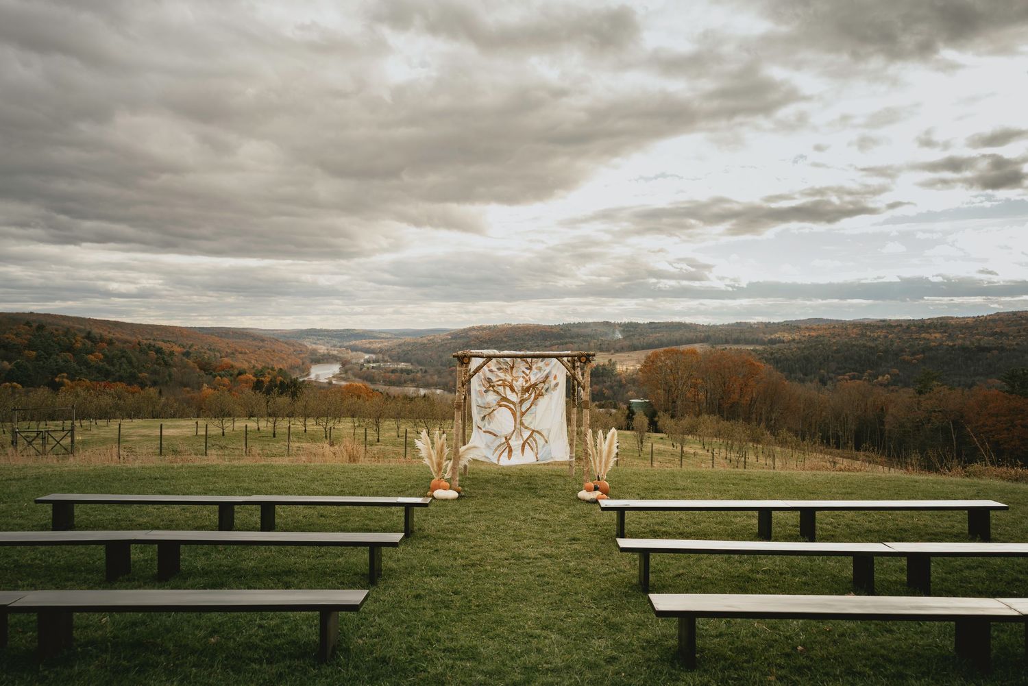 Outdoor wedding ceremony setup with wooden benches and decorative arch overlooking a scenic valley under dramatic cloudy skies.