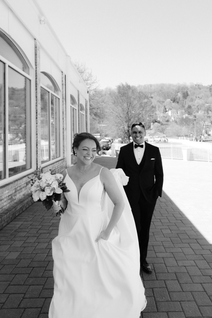 Black and white photo of a smiling bride in a white gown holding bouquet, groom in tuxedo walking behind her.