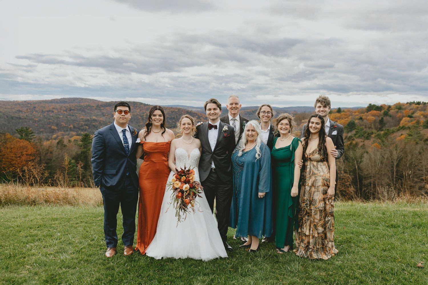 Wedding party gathers on grassy hillside with mountains and autumn colors in the distance.