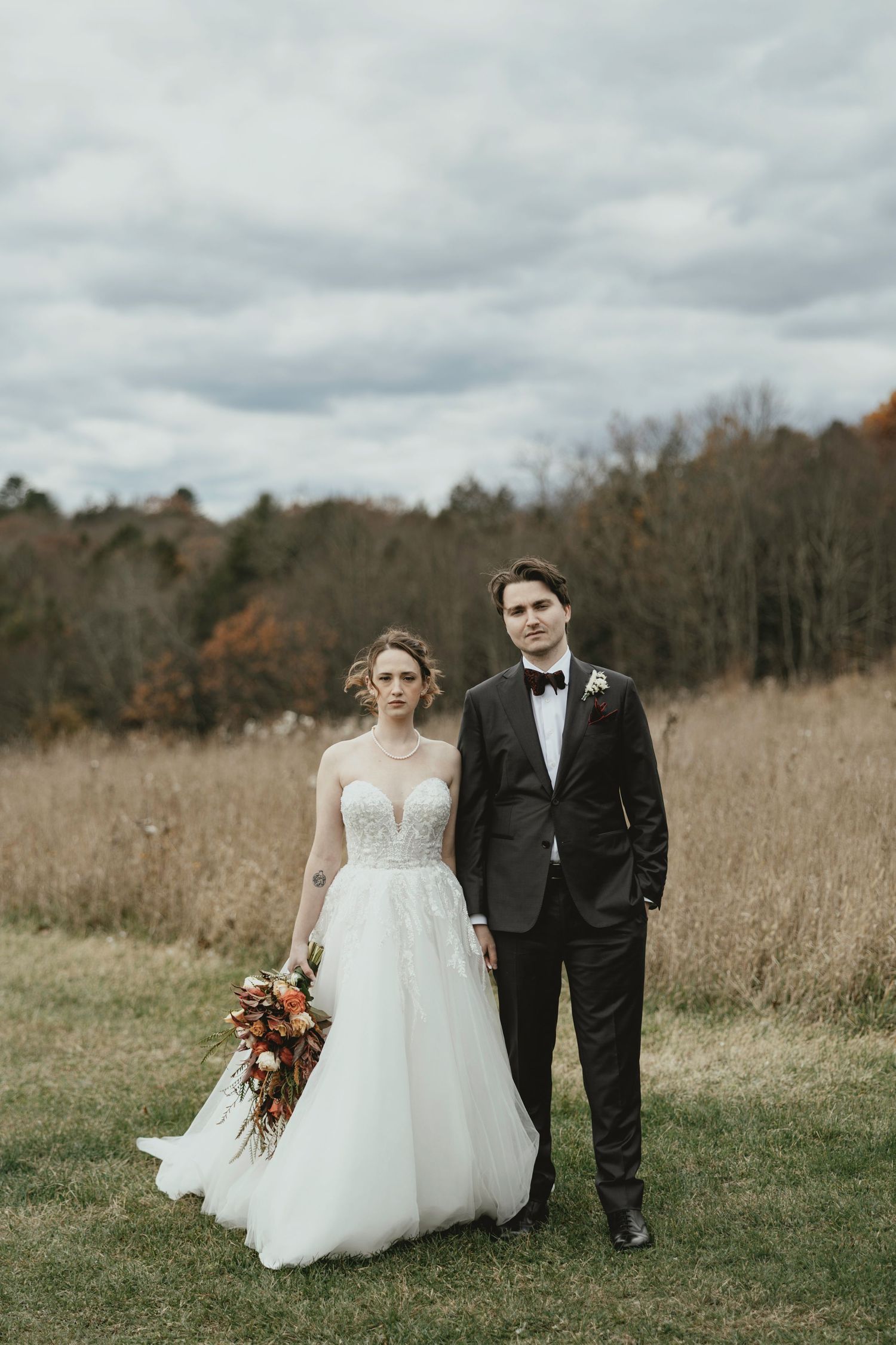 Couple in wedding attire walking through a scenic autumn field with tall grass and trees in the background.