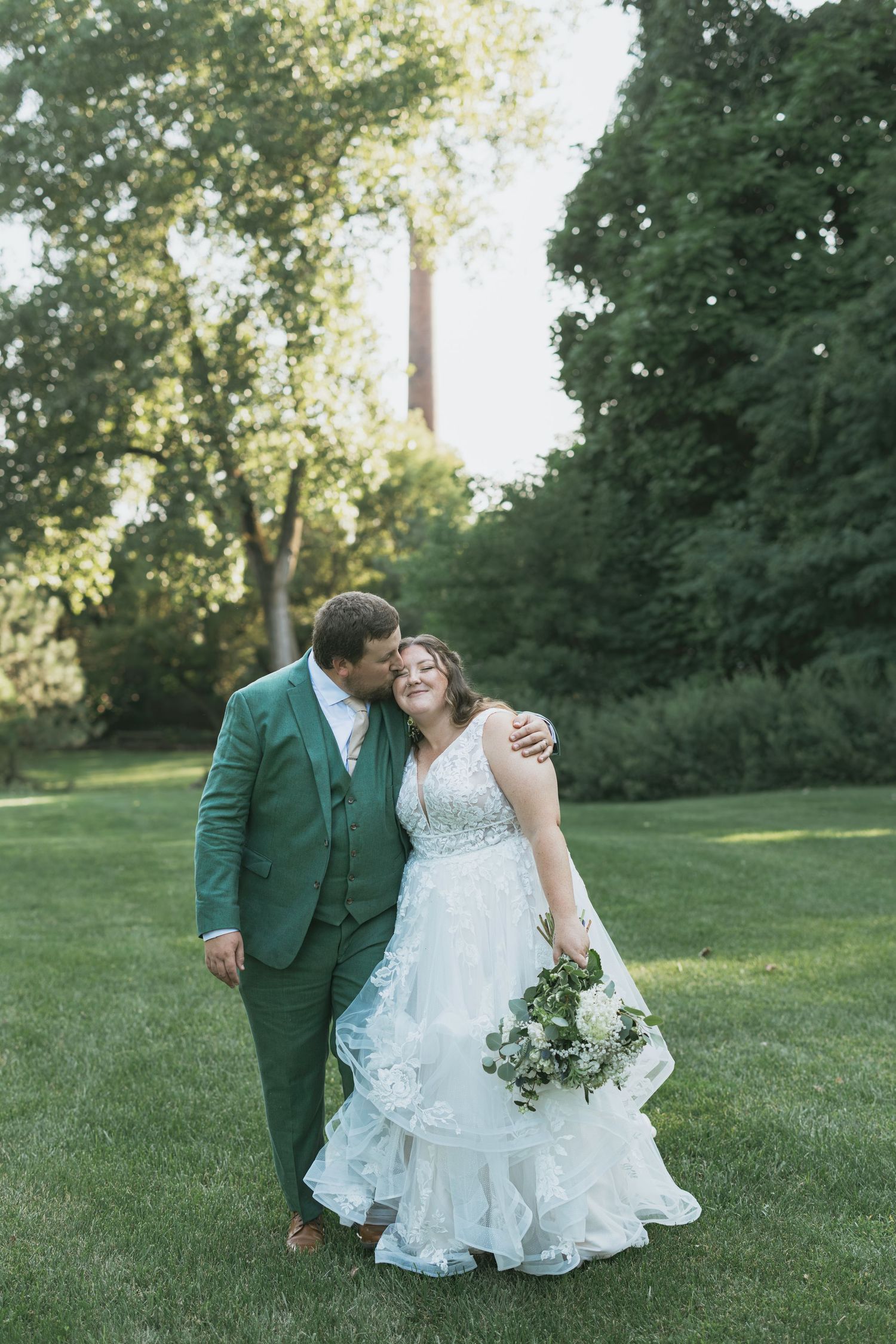 A couple in wedding attire share romantic moments on a green lawn with trees in the background during golden hour.