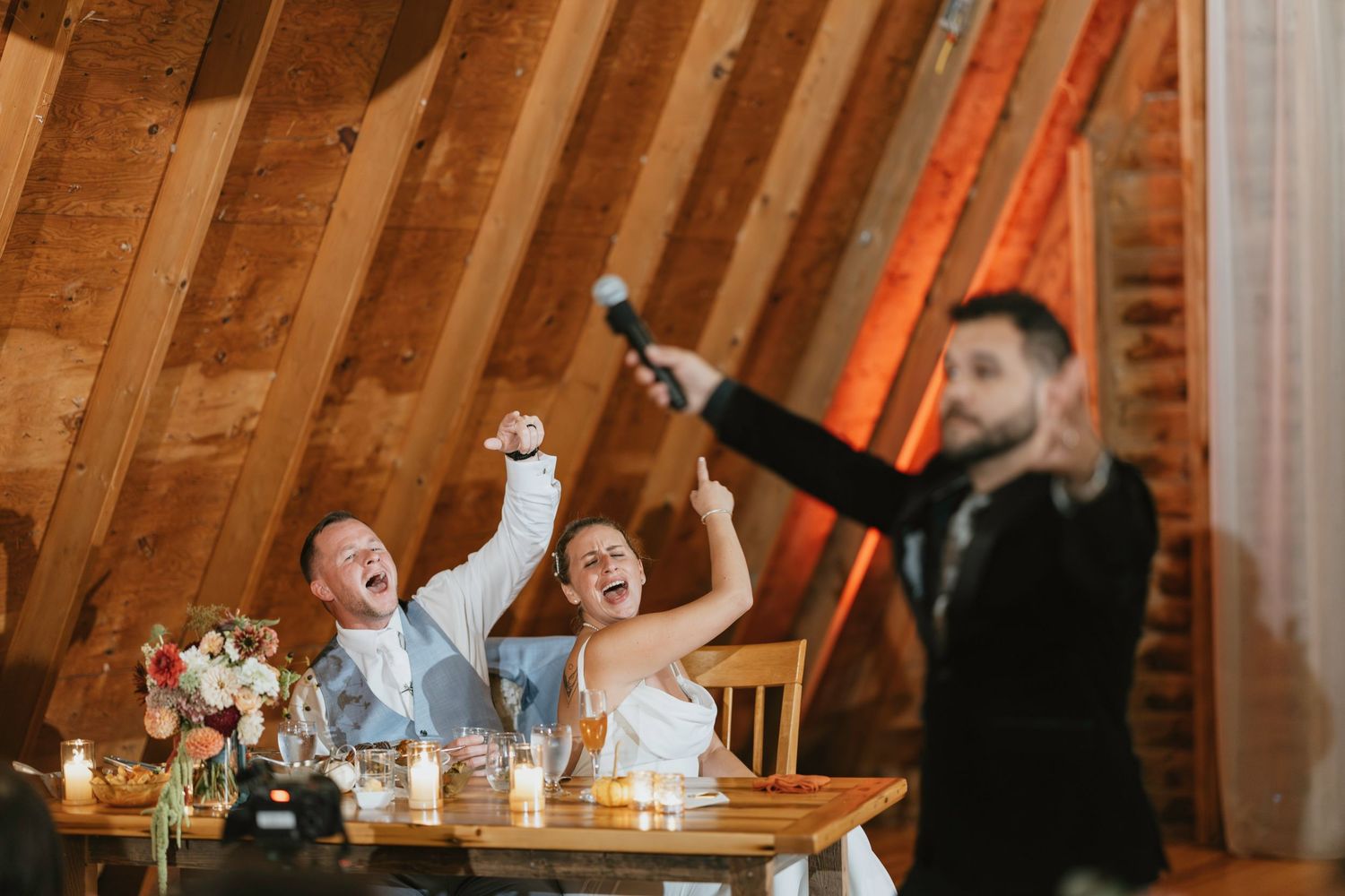 Wedding reception guests laughing and enjoying themselves at rustic wooden tables in barn venue with warm lighting.