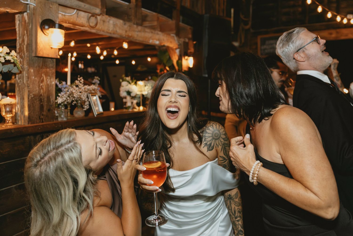 Wedding guests laugh and dance together under warm string lights at an evening reception celebration.