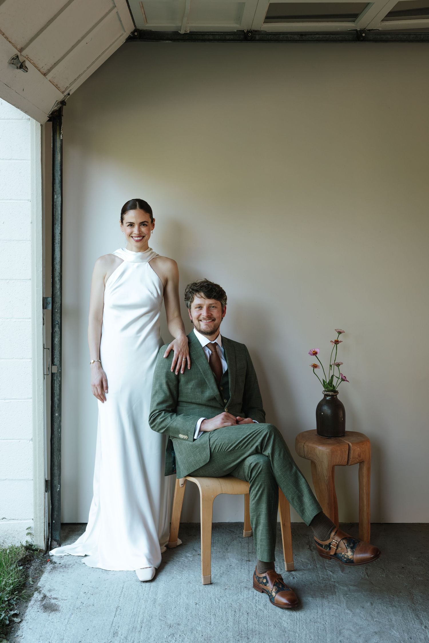 Bride in white halter gown stands beside groom seated in chair wearing green suit, posing for wedding portrait.