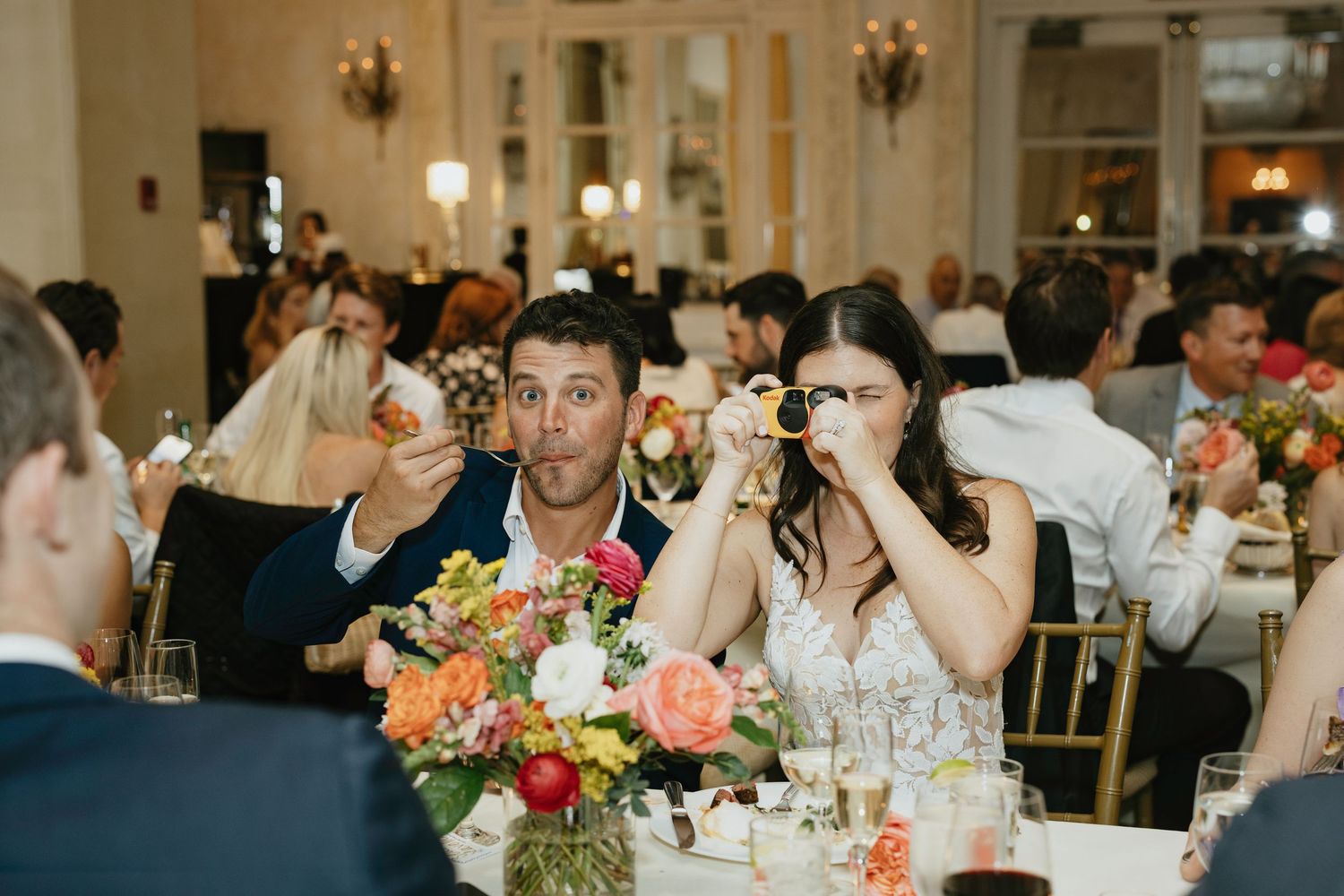 Wedding reception guests gathered at elegantly decorated tables with floral centerpieces in a warmly lit ballroom.