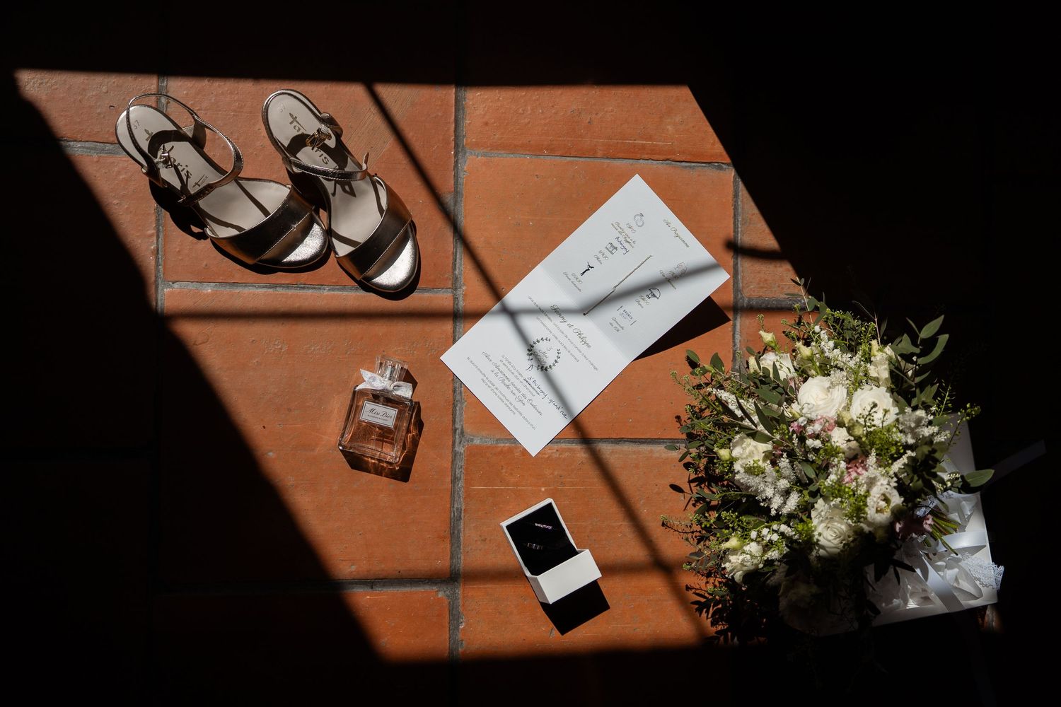 Wedding accessories including sandals, invitation, perfume bottle, and bouquet arranged on terracotta tiles in sunlight.