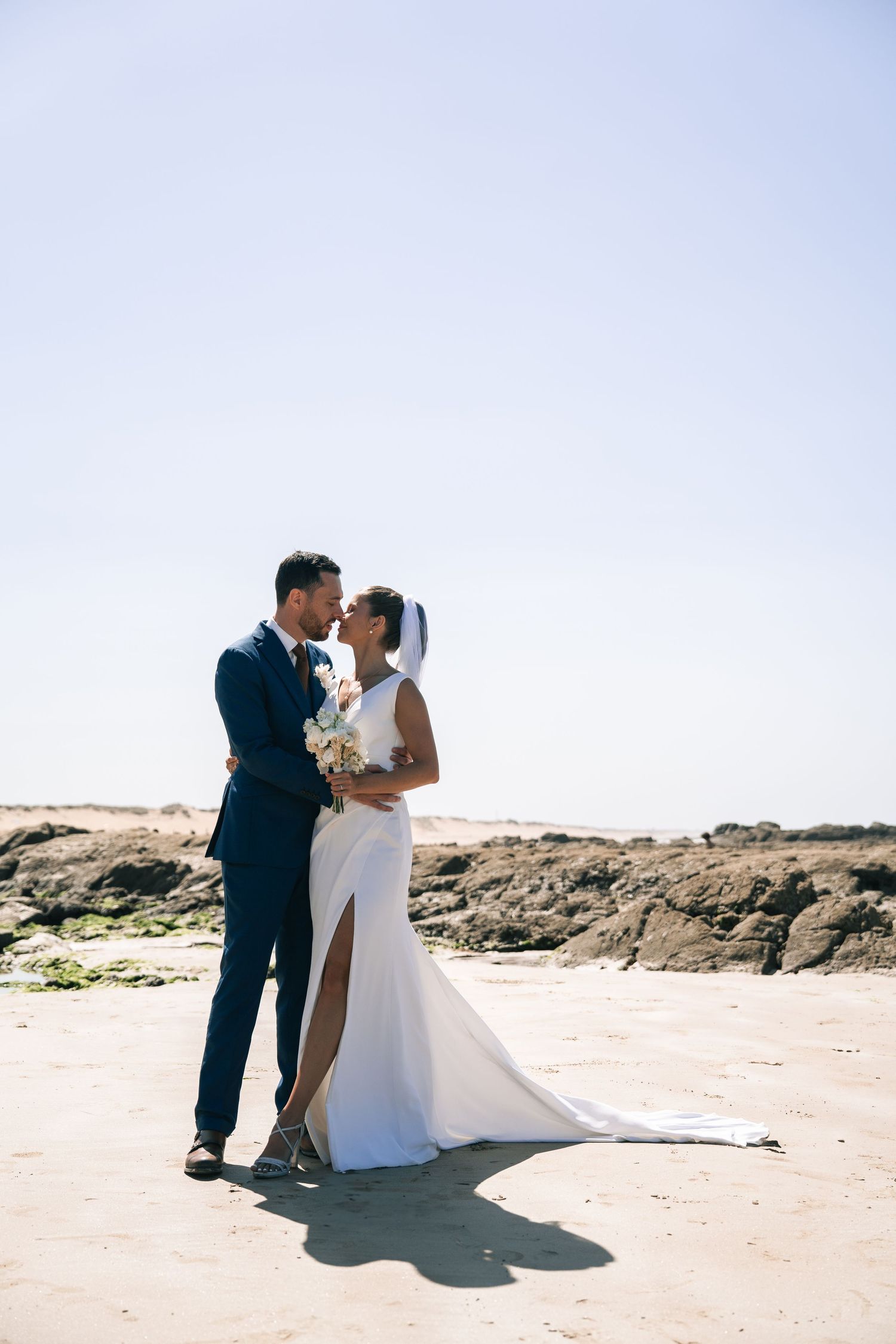 A romantic wedding couple shares intimate moments on a sunny beach with rocky cliffs in the background.