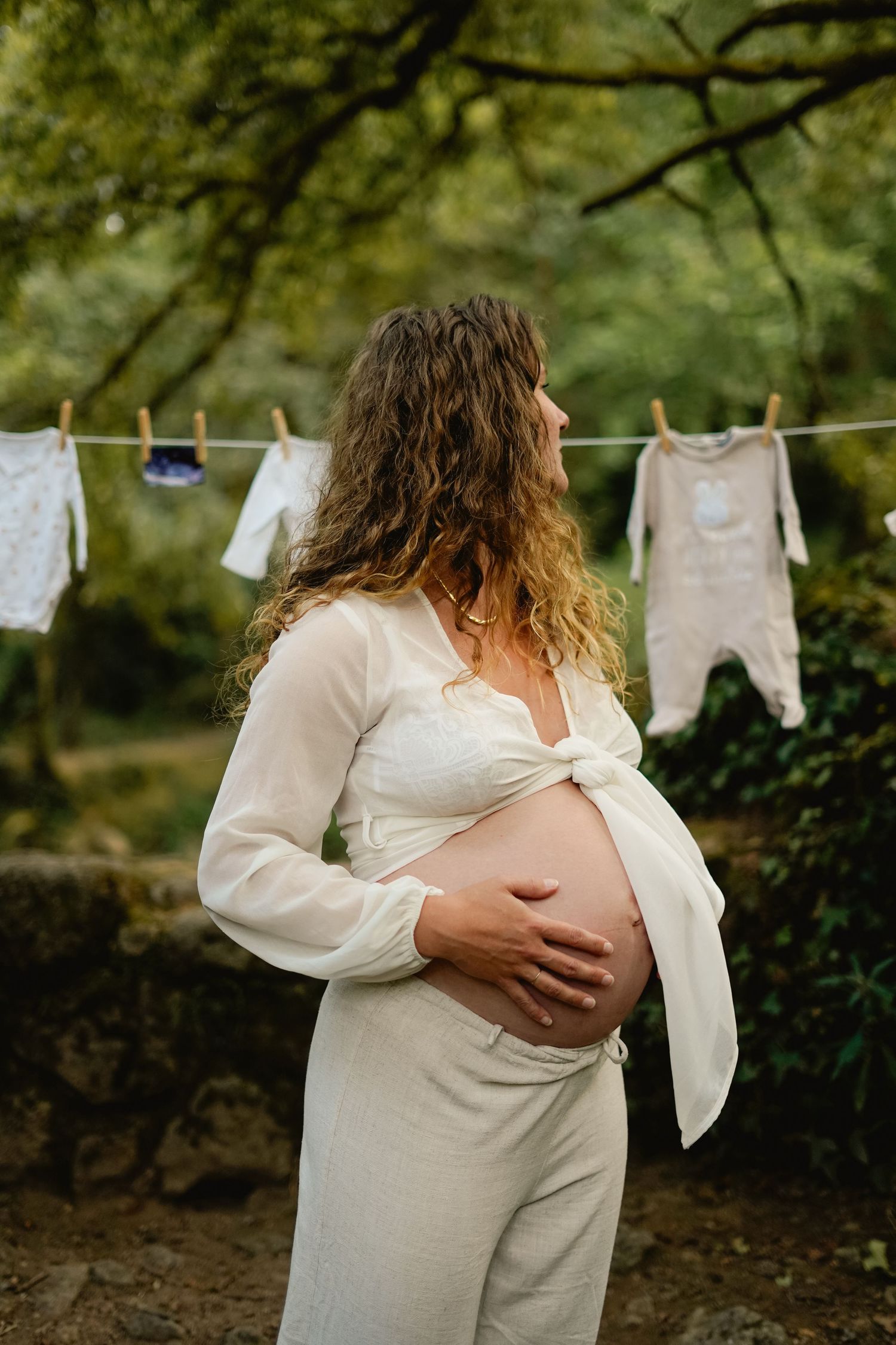 Expectant mother in white dress standing near clothesline with baby clothes in garden setting.