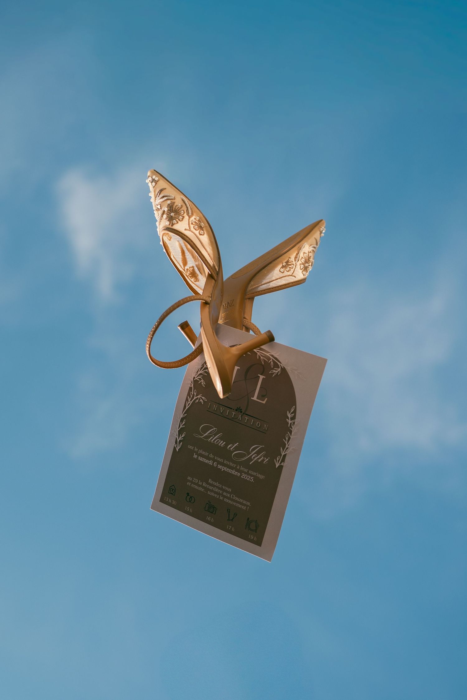 A wedding invitation card floats against a bright blue sky with fluffy white clouds in the background.