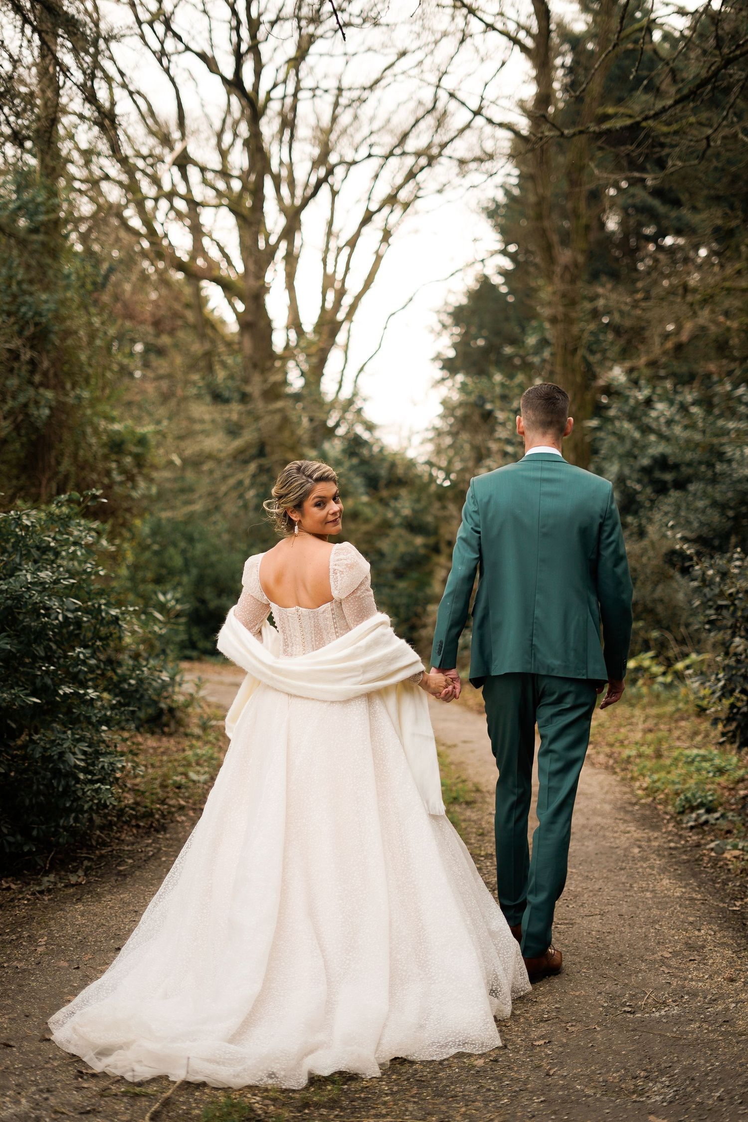 A couple walking down a wooded path in their wedding attire.