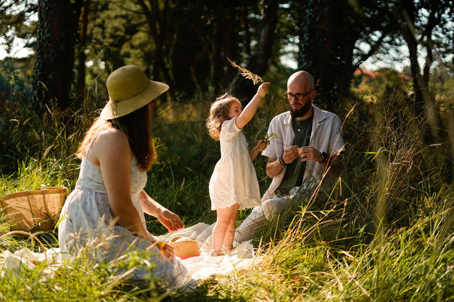 Family enjoying a sunny summer picnic in tall grass with trees in background.