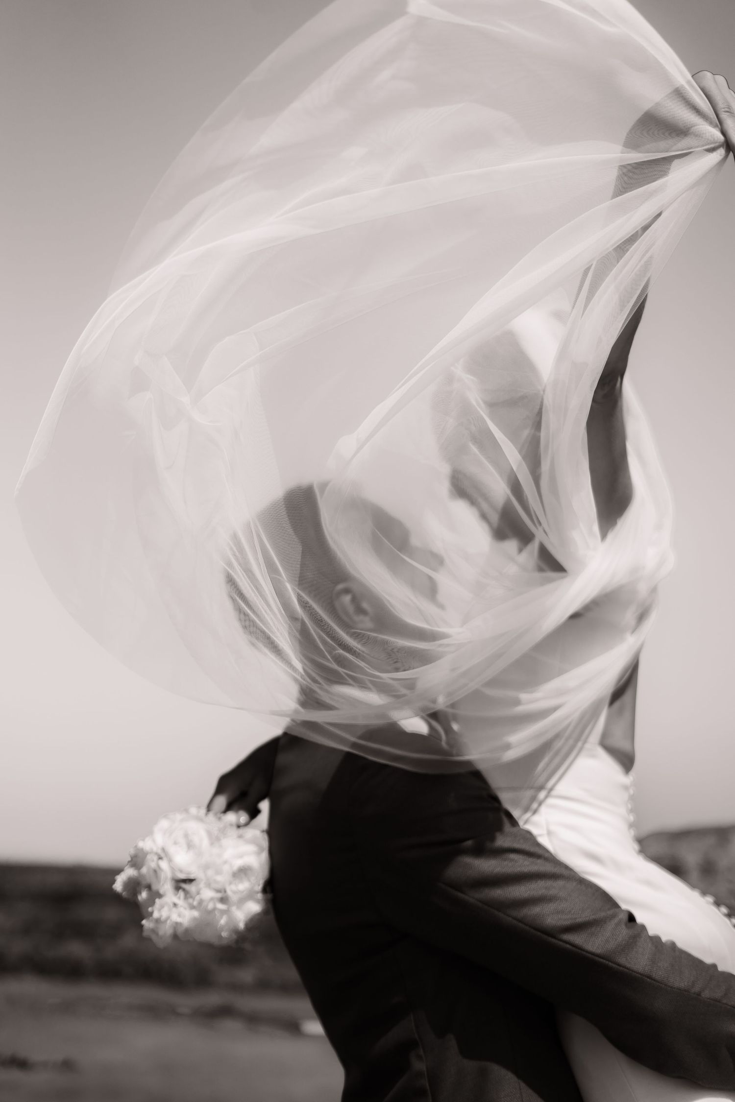 A flowing white wedding veil billows dramatically in the wind against a monochrome background.