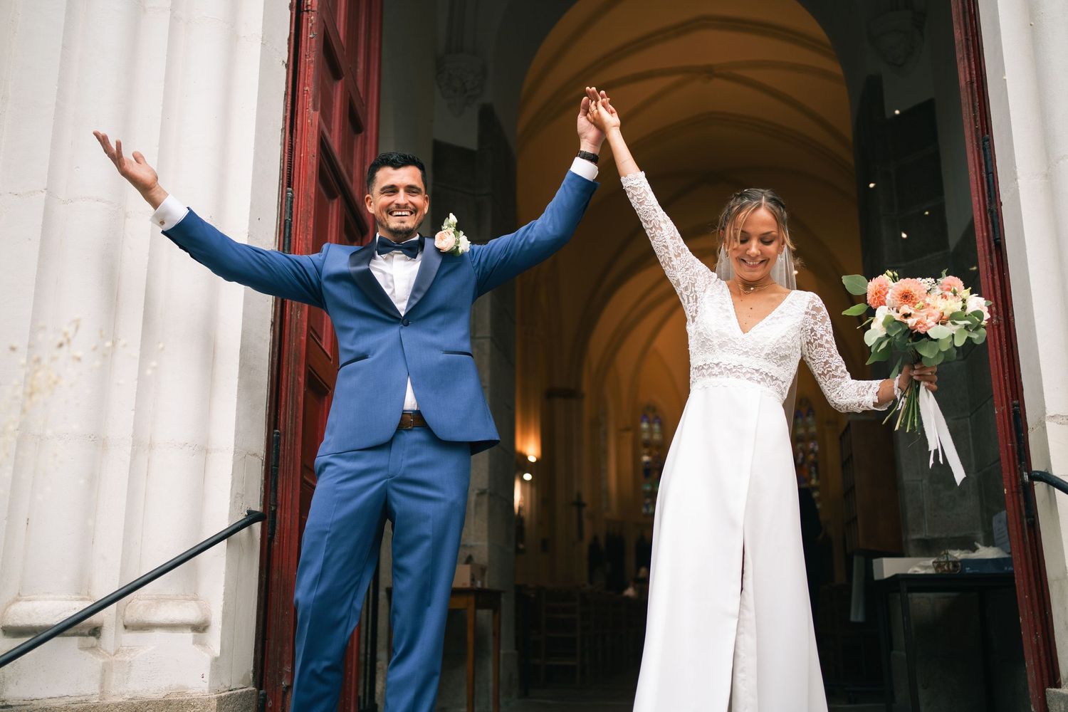 Newlywed couple and wedding party celebrate with raised arms in front of church entrance.
