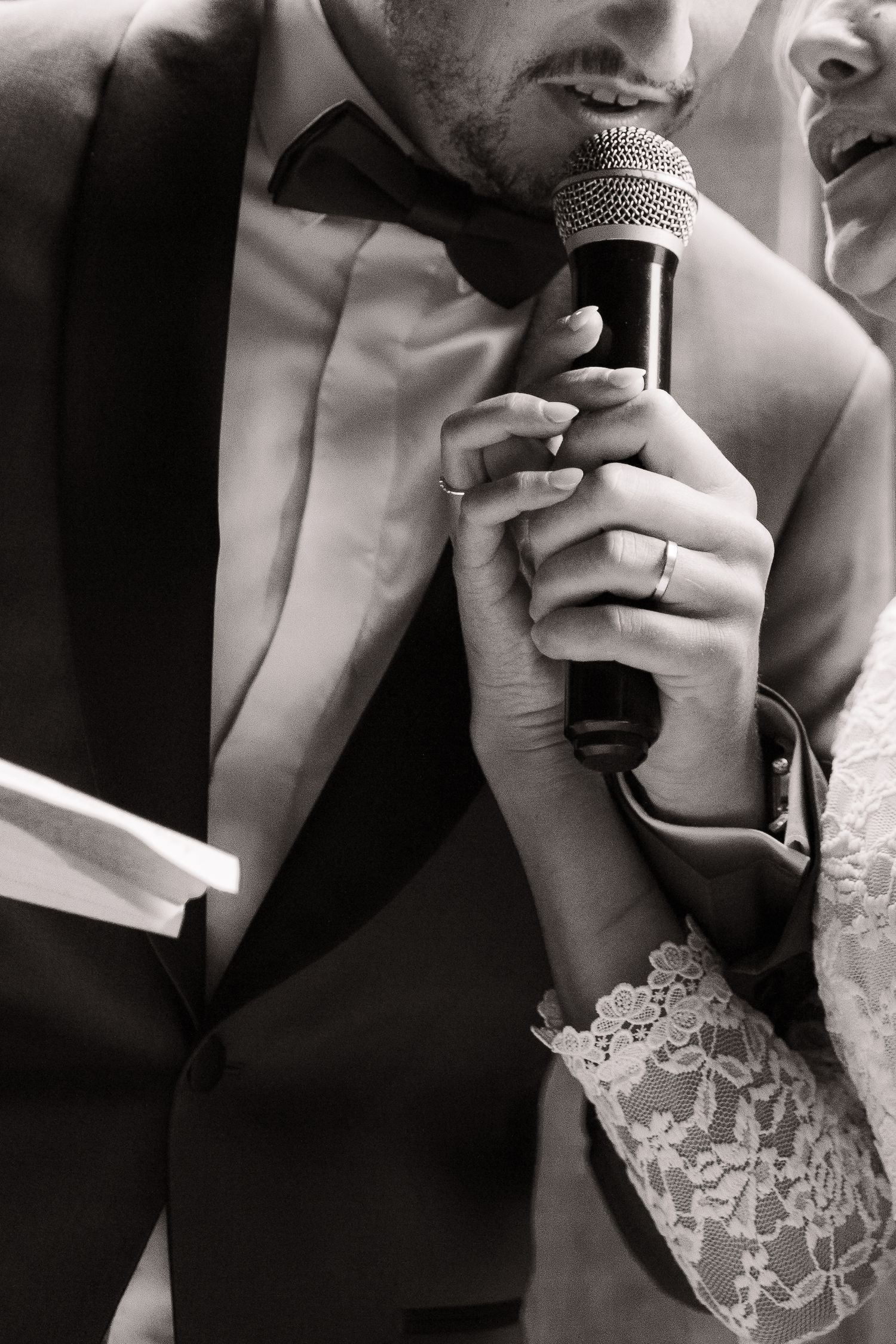 A close-up black and white photo of a couple sharing a microphone, with lace sleeve detail visible.