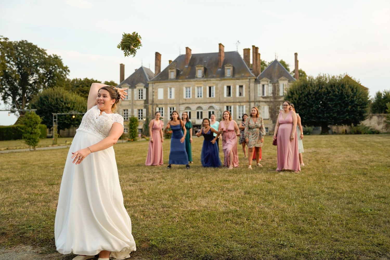 Elegant wedding party poses on the lawn in front of a majestic French chateau with a white-gowned figure in the foreground.