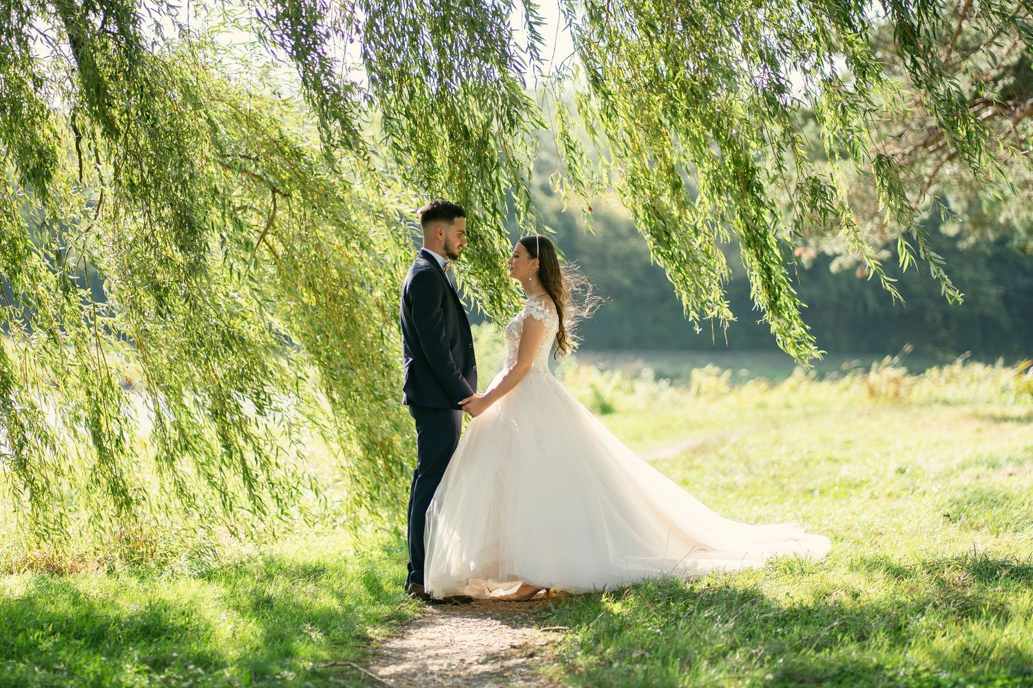 A couple in wedding attire walks along a scenic path under weeping willow trees on a sunny day.