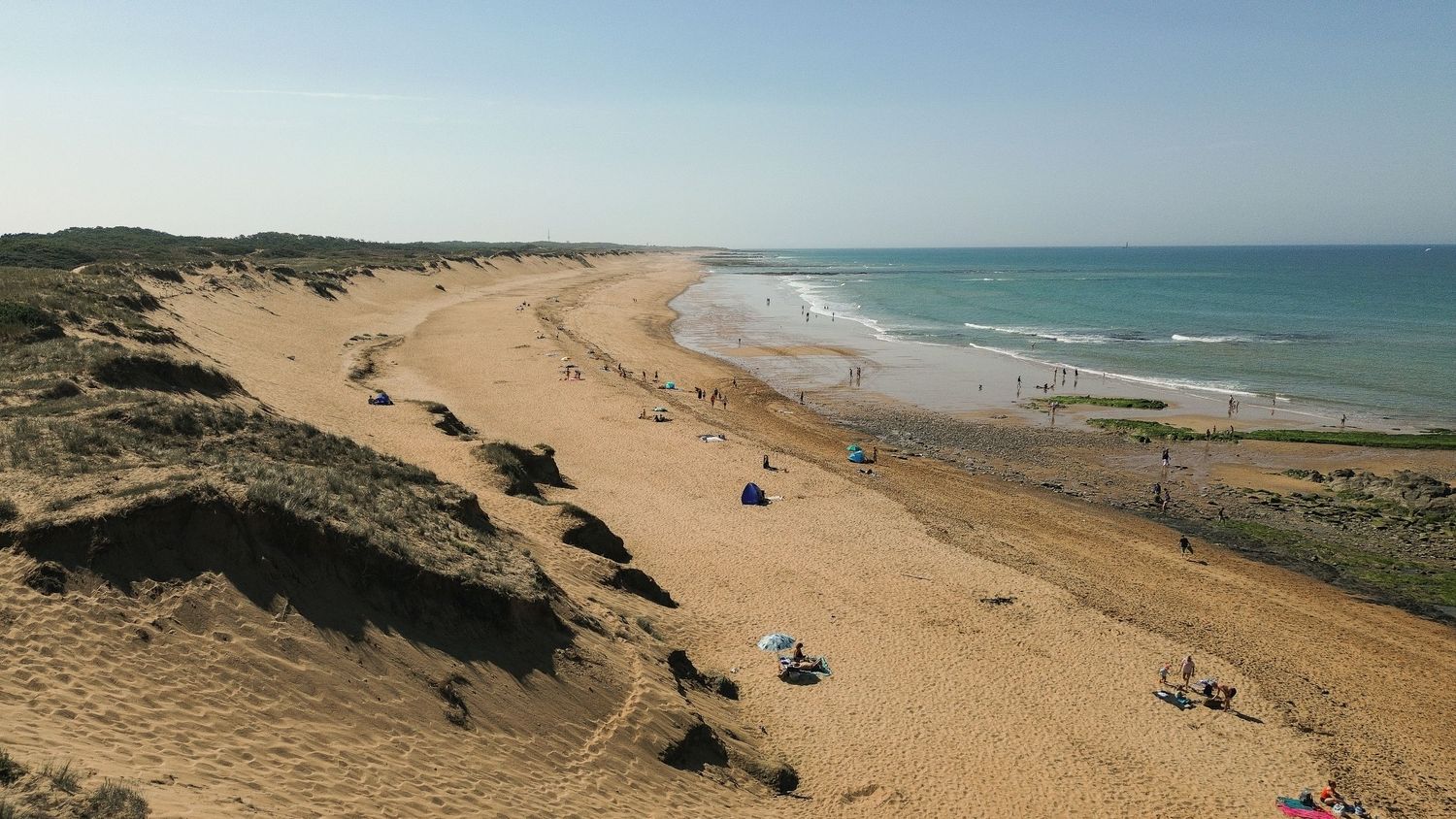 Panoramic view of sandy beach coastline with ocean waves and dunes stretching along the horizon under blue skies.