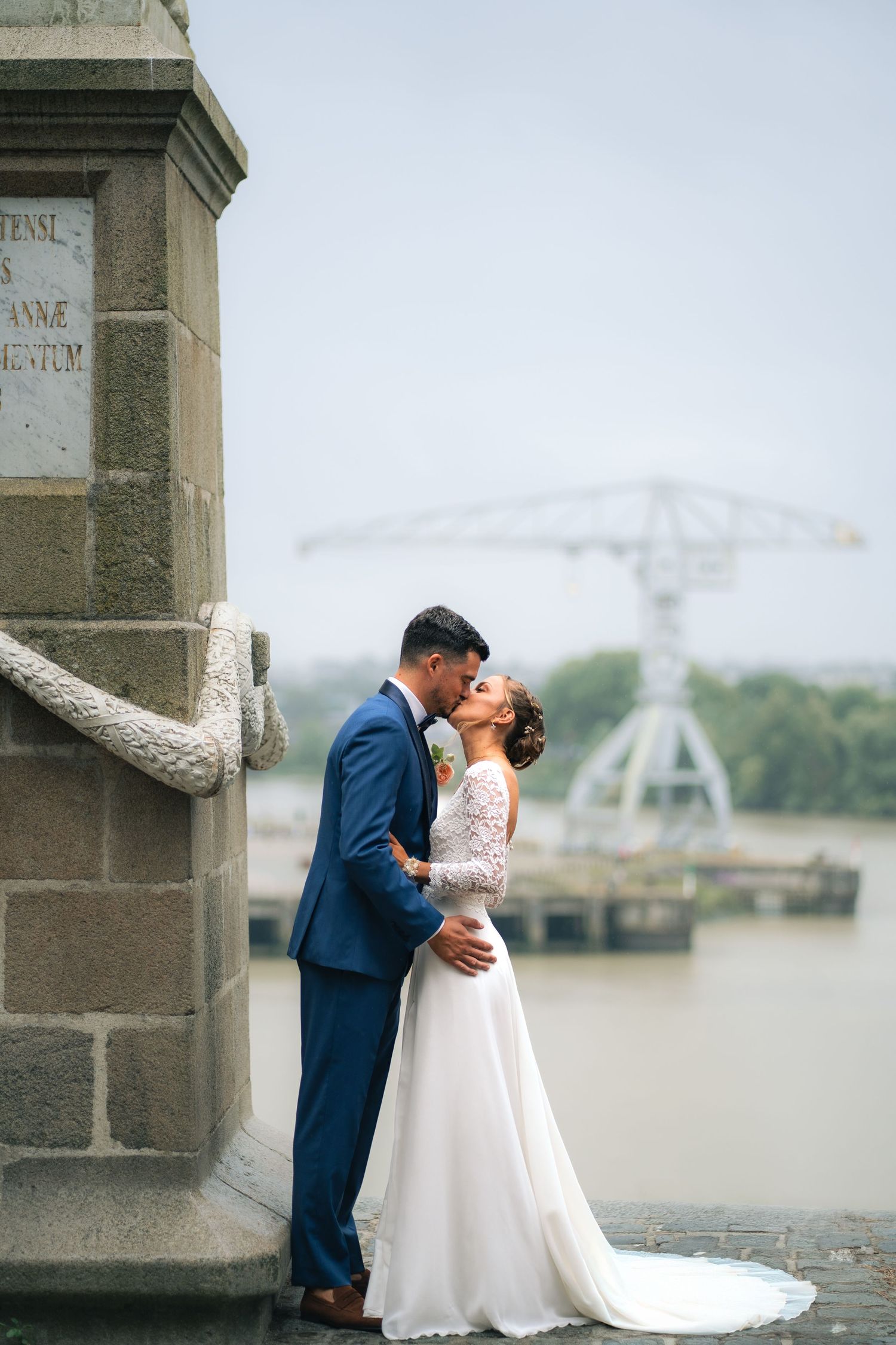 A romantic wedding couple embraces by a riverfront stone pillar on a misty day.