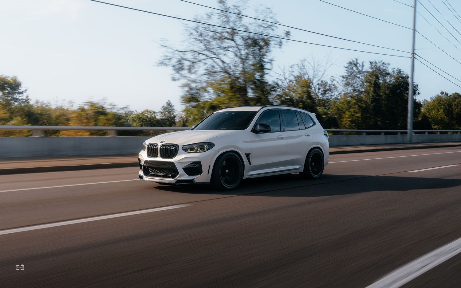 White BMW X3 SUV with custom wheels driving on a highway with power lines and trees in the background.