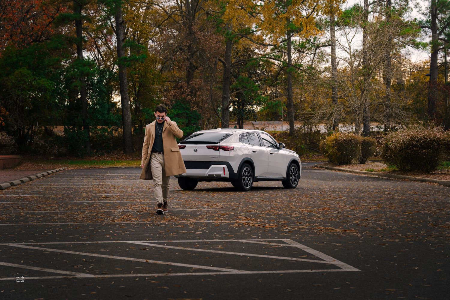Autumn leaves scatter across an empty parking lot with trees and cars visible in the background.