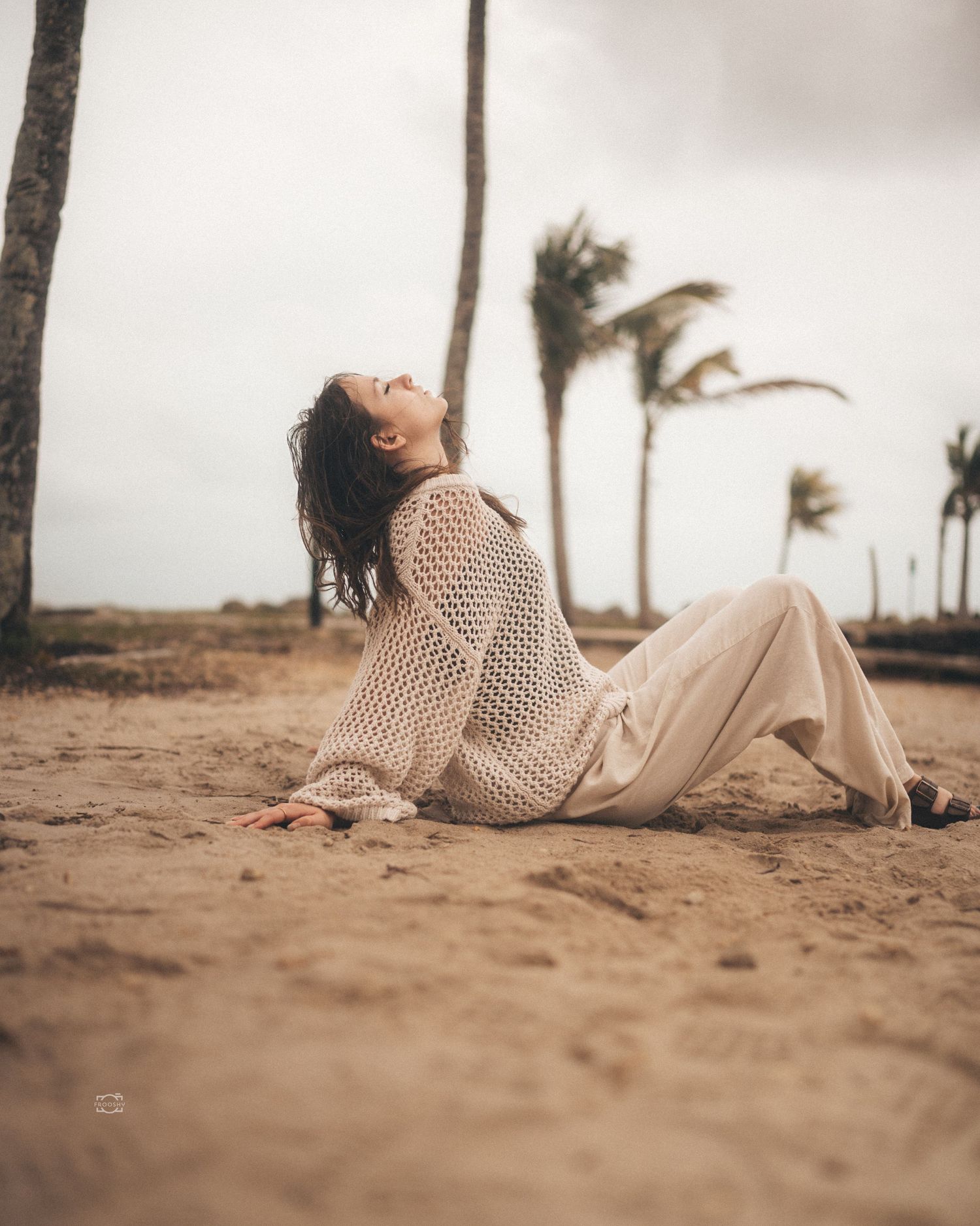 A person in a beige sweater lies on a sandy beach with palm trees swaying in the wind on a cloudy day.