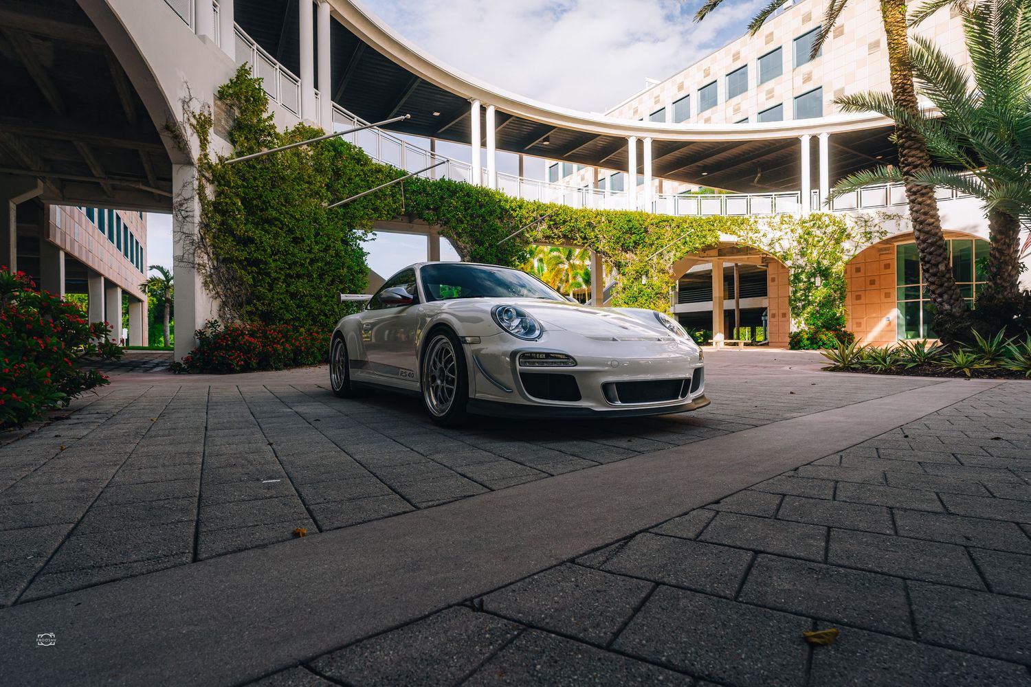 A silver Porsche 911 GT3 parked in a curved modern architectural setting with palm trees and greenery.