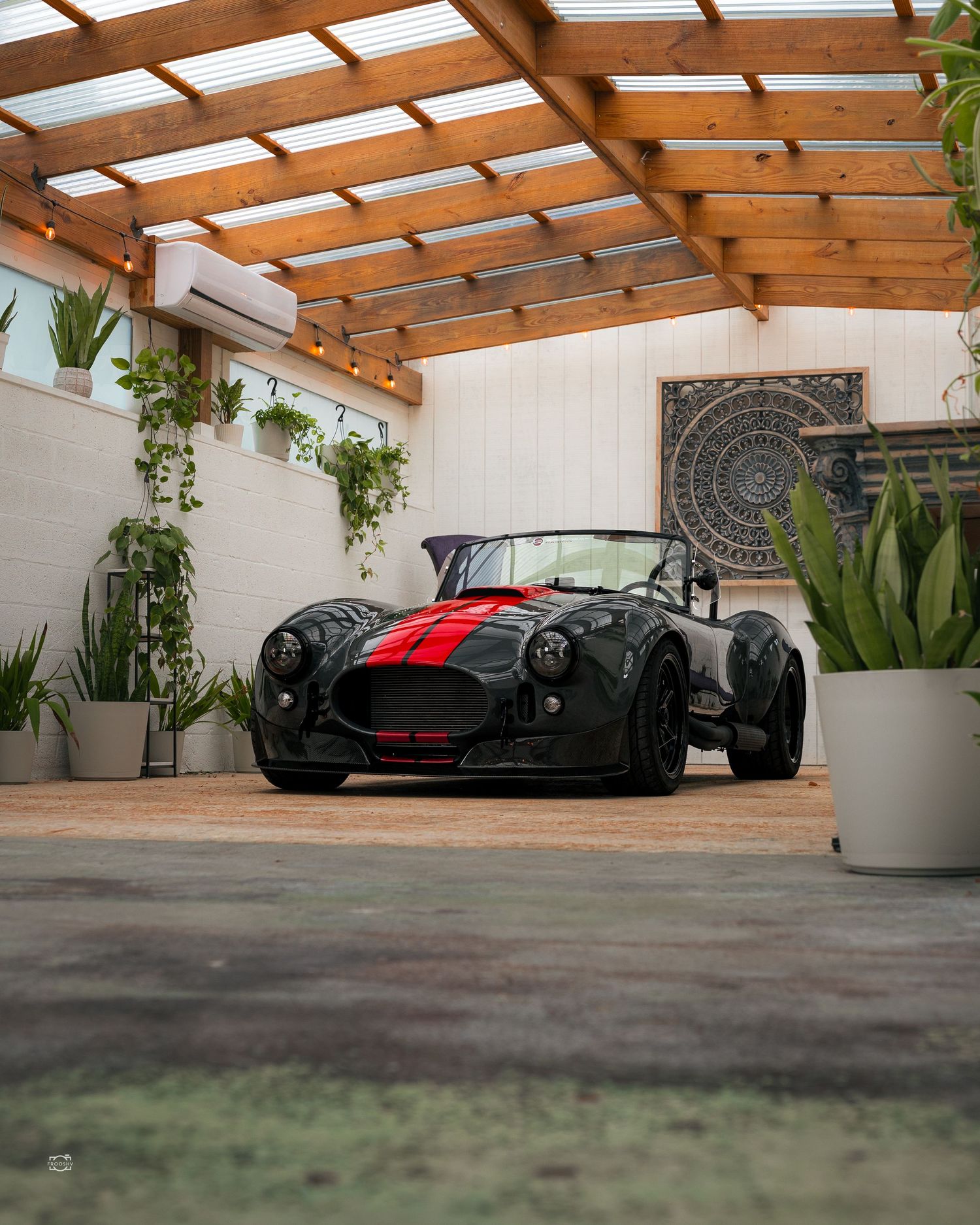 A black vintage Cobra roadster with red racing stripes parked under a wooden pergola with hanging plants and modern decor.