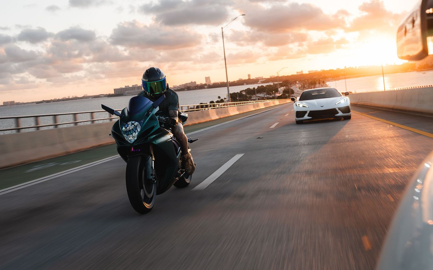 A motorcycle rider leans into a curve on a coastal highway during a dramatic orange sunset.