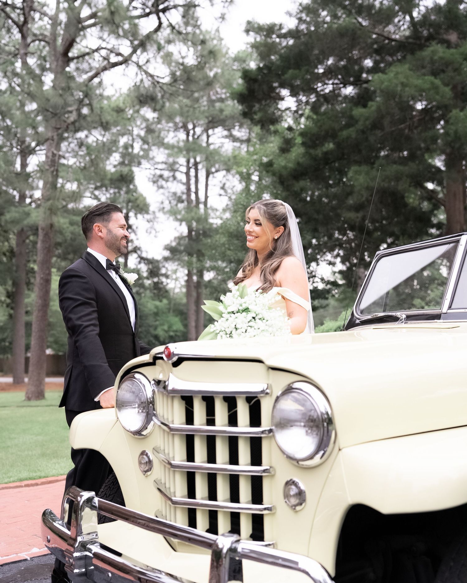 Newlyweds pose with a classic cream-colored vintage car on their wedding day against a backdrop of tall pine trees.