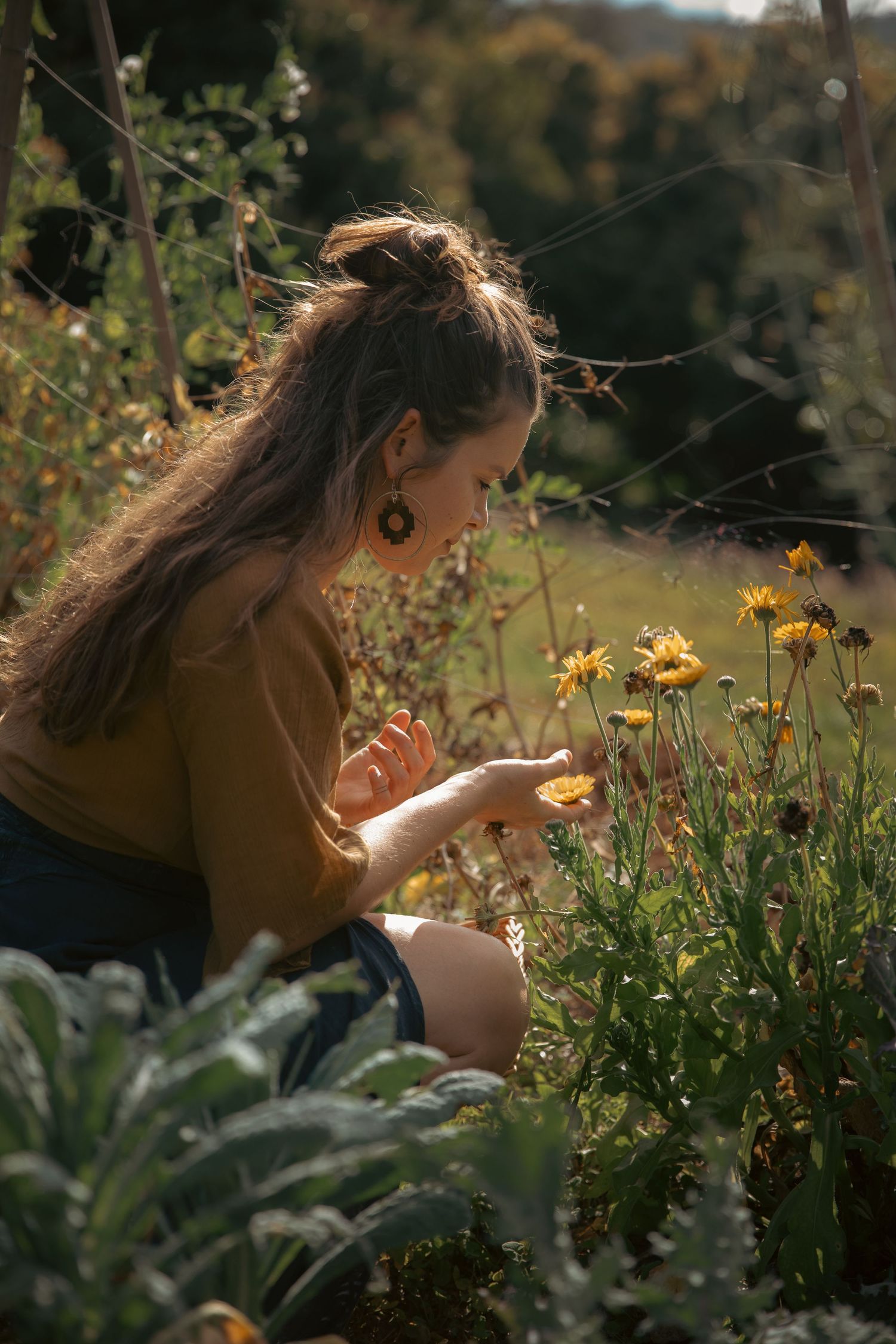 Young woman with long hair crouching in a sunlit garden carefully examining yellow wildflowers.
