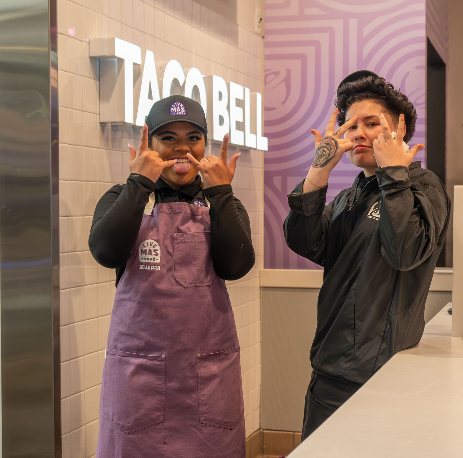 Two Taco Bell employees in purple aprons posing with hand gestures in front of the Taco Bell sign.