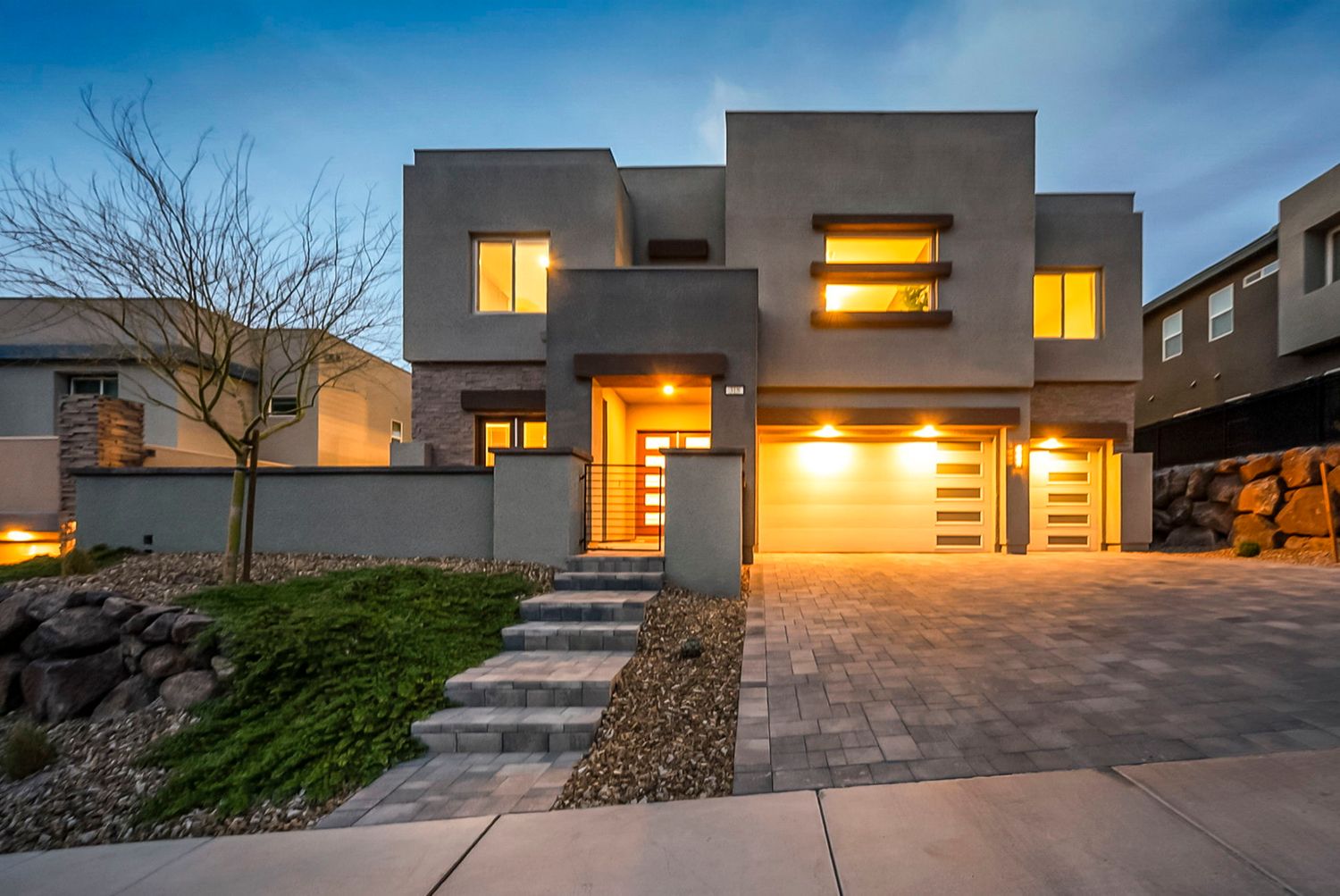 Modern two-story stucco home with warm glowing windows, stone steps, and a two-car garage at dusk.