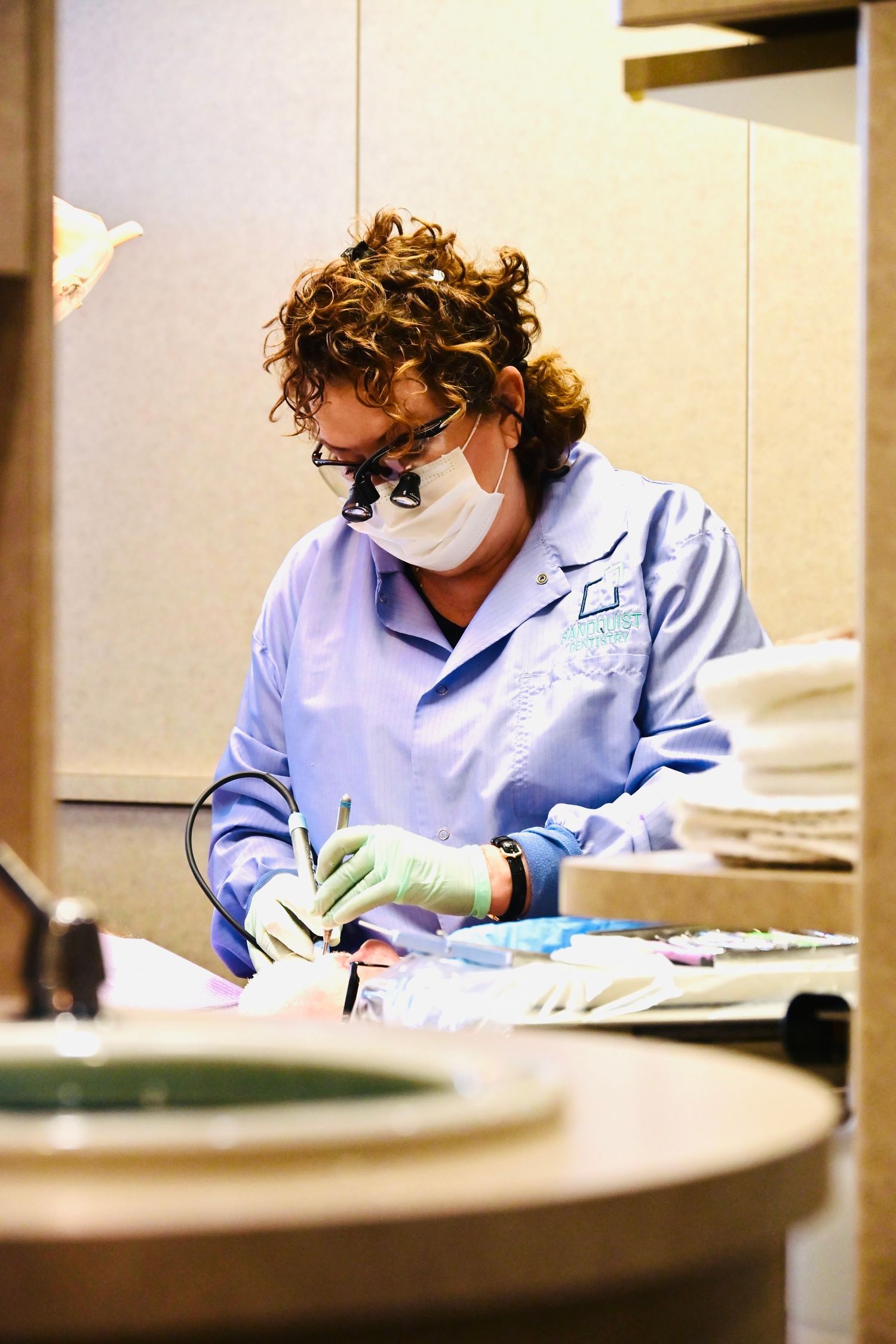 Female dental hygienist with curly hair wearing loupes and mask performs dental procedure on patient.