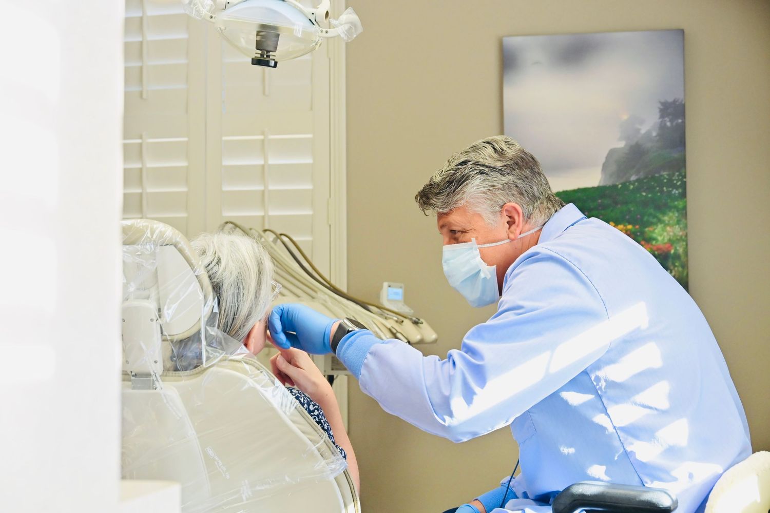 Male dentist in blue scrubs examines seated patient in bright dental treatment room.