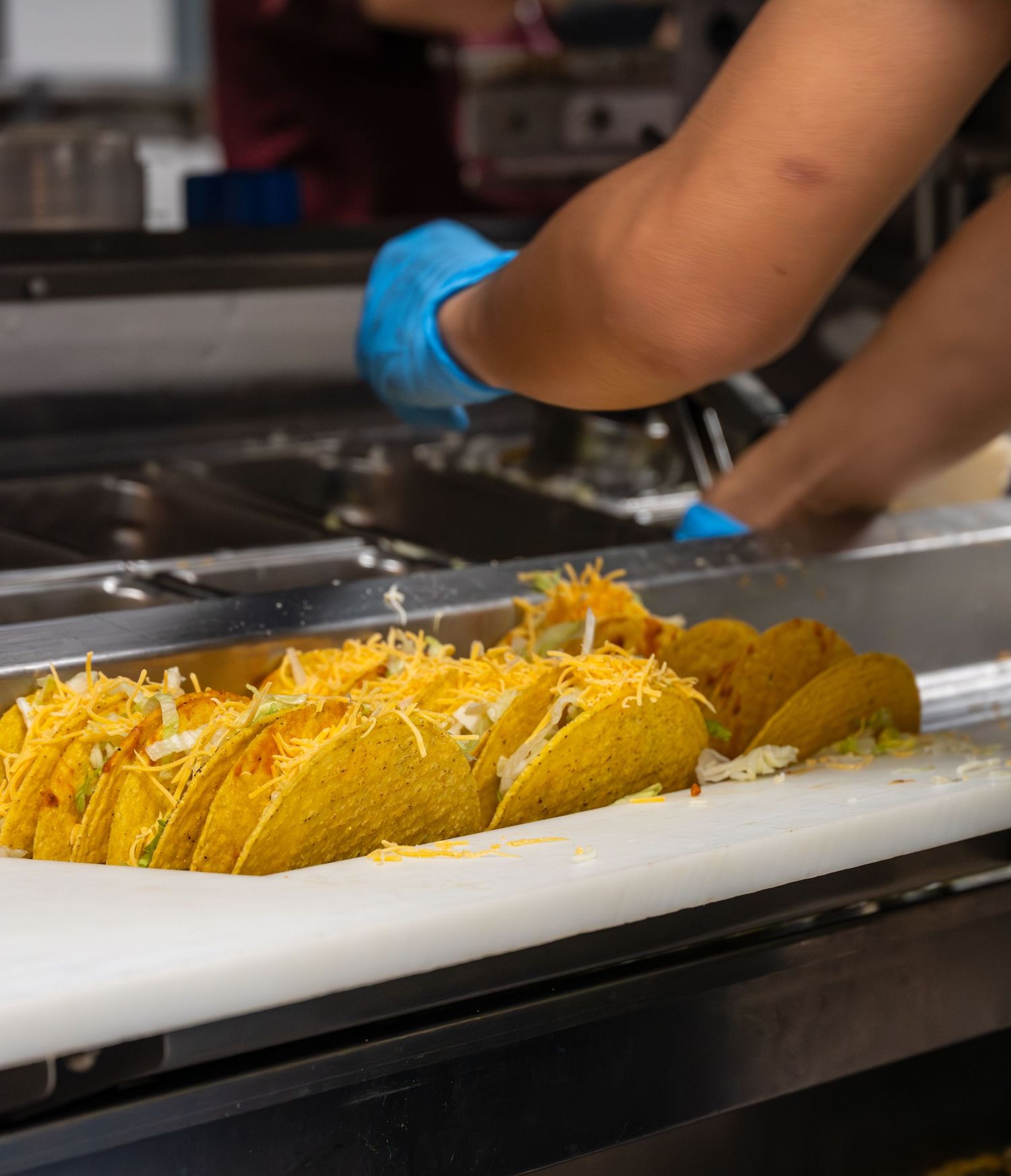 Taco Bell employee in blue gloves preparing multiple crunchy tacos with cheese on a white tray.