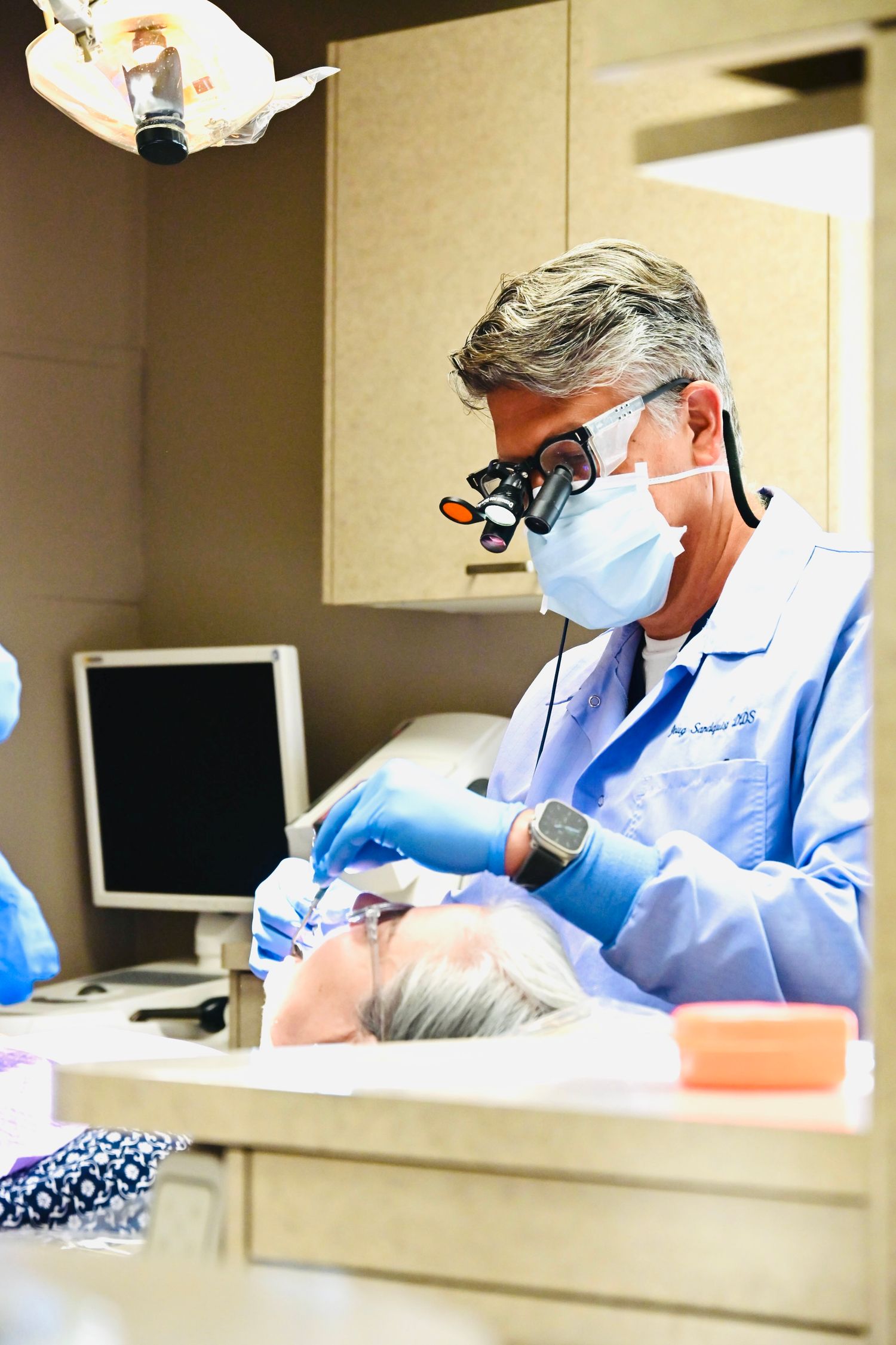 Male dentist with loupes and mask works on patient while assistant observes in dental operatory.