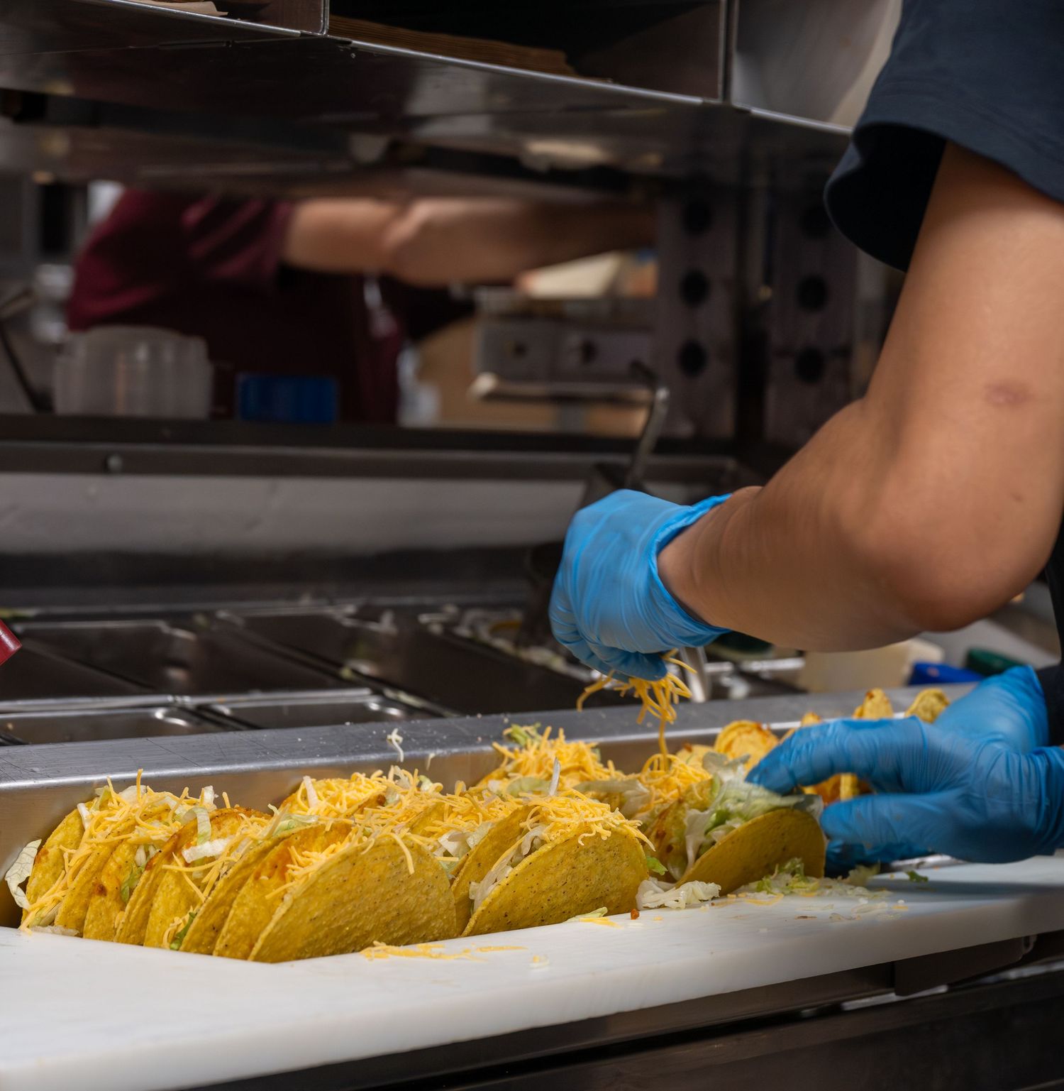 Taco Bell kitchen worker in blue gloves assembling a row of crunchy tacos on a tray.