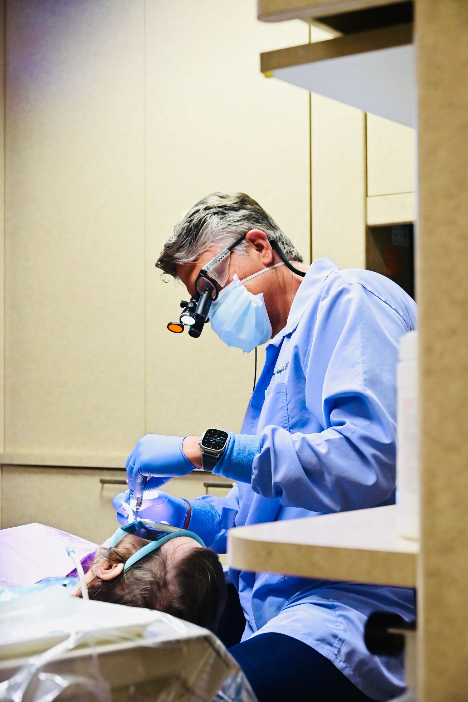 Dentist wearing magnifying loupes and mask performs detailed dental procedure on reclined patient.