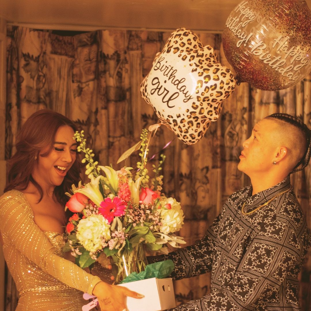 Man presenting birthday girl with flowers and leopard print balloon at indoor celebration.