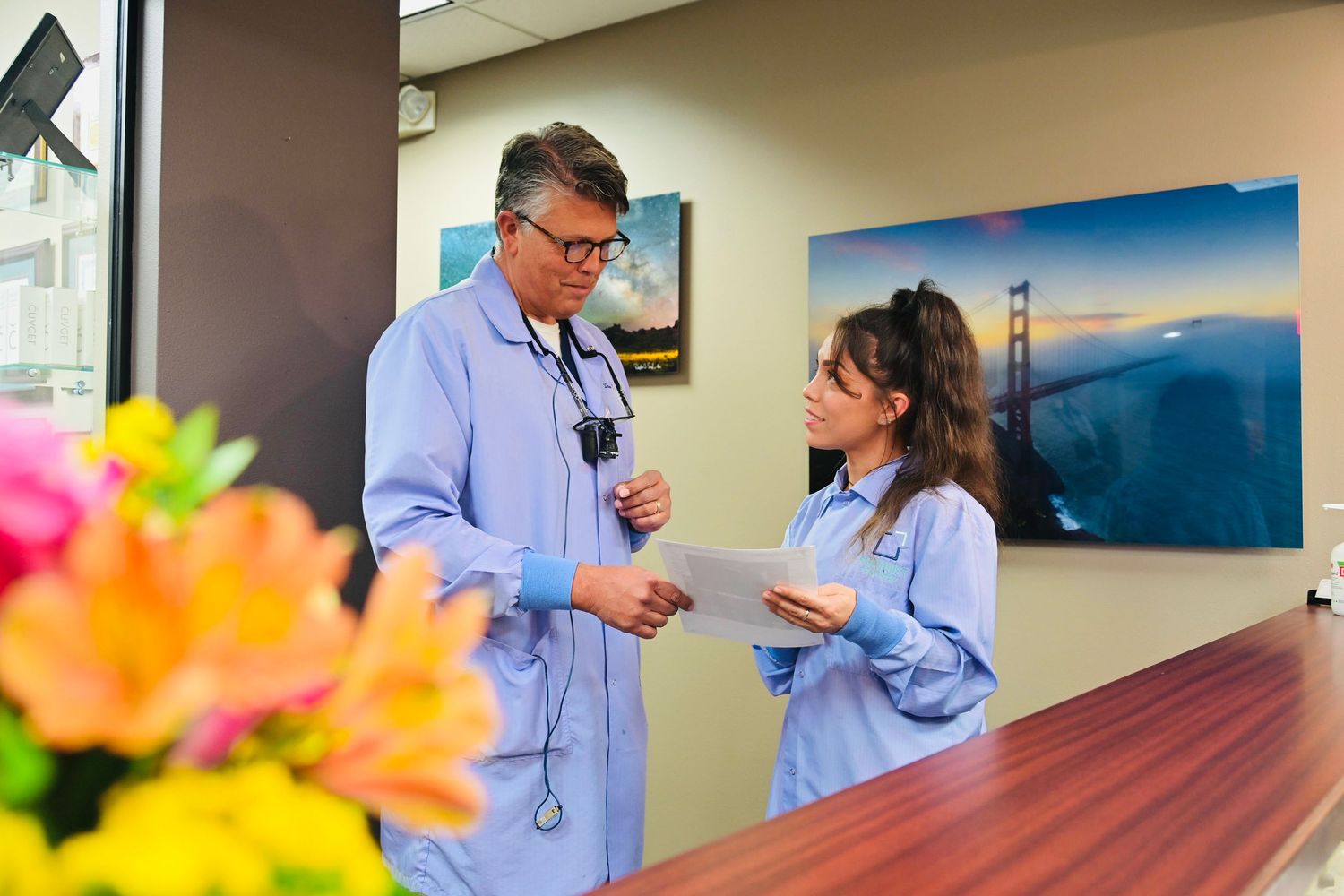 Dentist and female staff member discuss paperwork at front reception desk of dental practice.