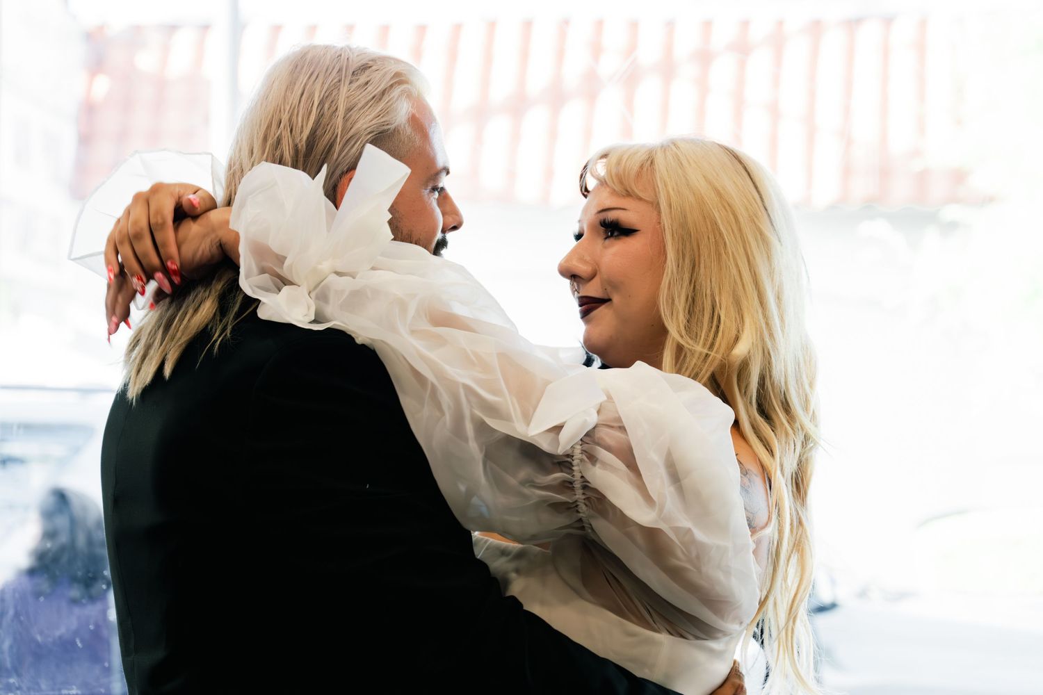 A joyful bride in a white sheer top embraces a guest with long blonde hair at a wedding celebration.