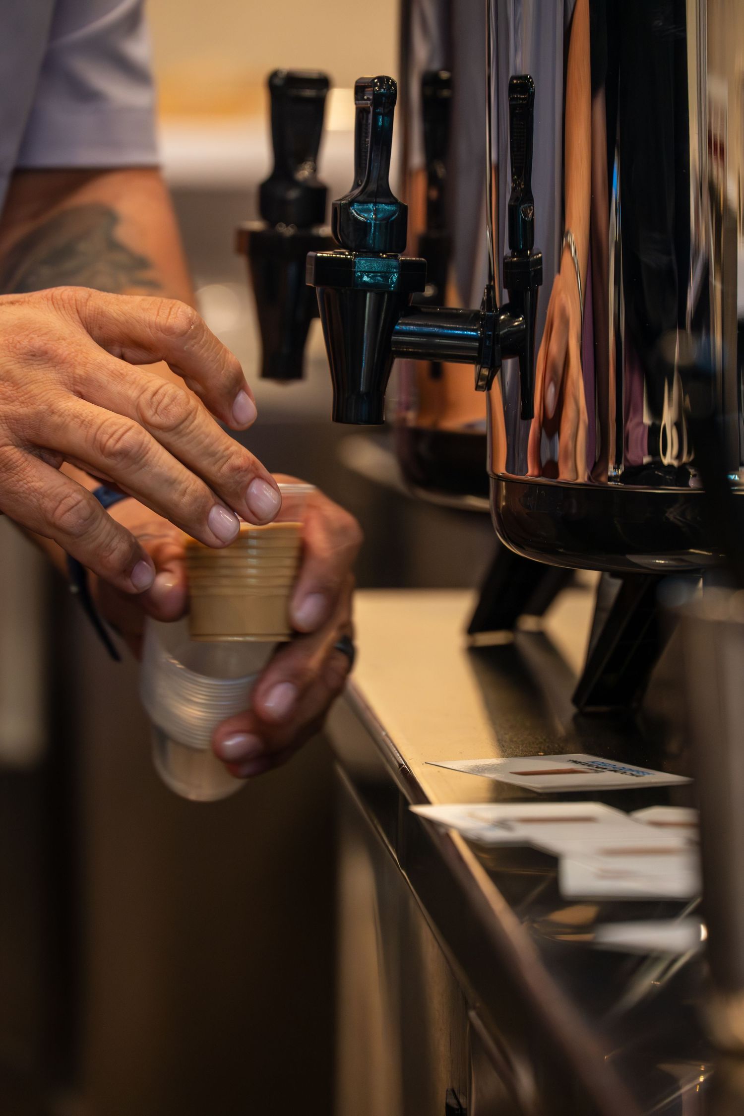 Tattooed bartender filling a small cup at sleek black beverage dispensers on a bar counter.