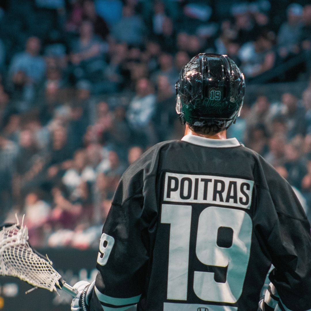 Lacrosse player number 19 Poitras standing on arena floor with stick in front of crowd.