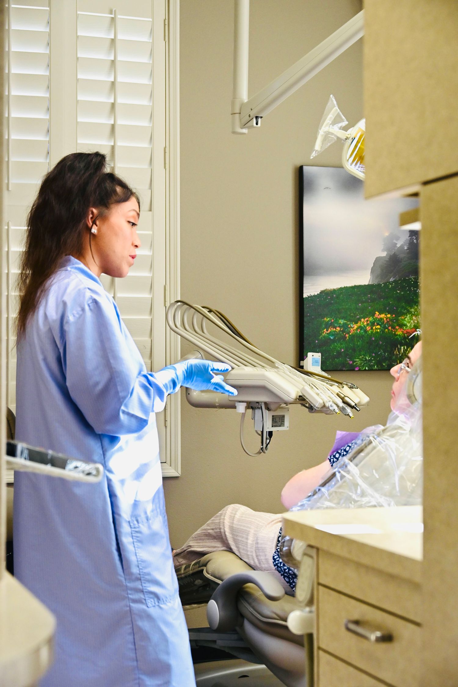 Female dental assistant in blue scrubs adjusts equipment while speaking with patient in dental chair.