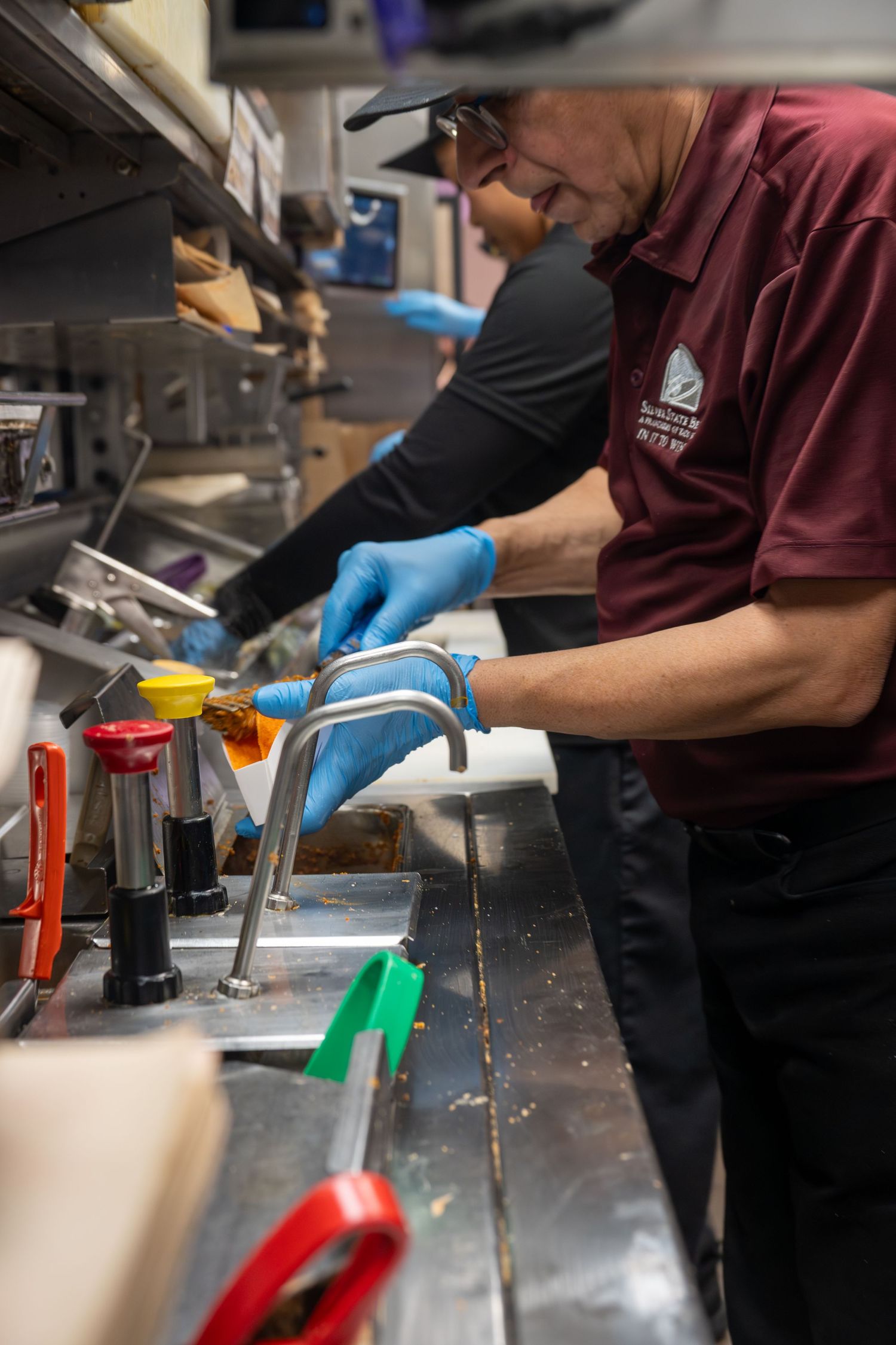 Taco Bell kitchen employee in maroon shirt and blue gloves assembling food at the prep station.