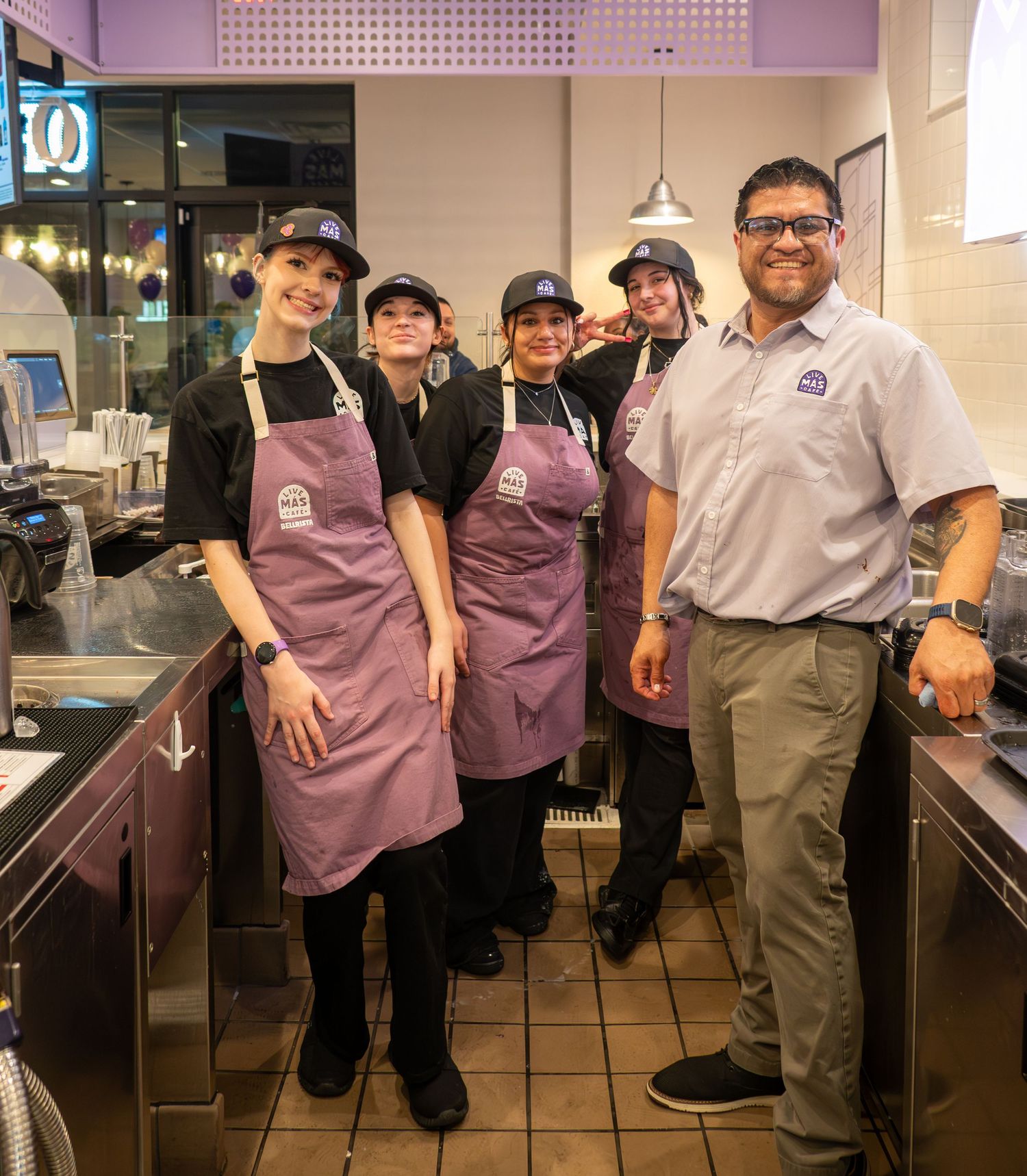 Group of four Taco Bell employees in purple aprons posing with a manager in the kitchen.