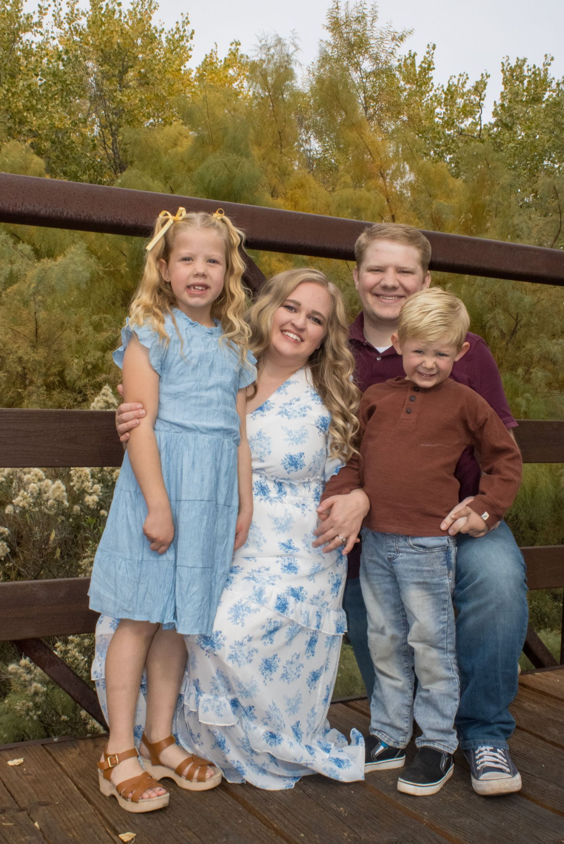 Happy family of four posing on a wooden bridge surrounded by autumn trees, wearing blue and casual outfits.